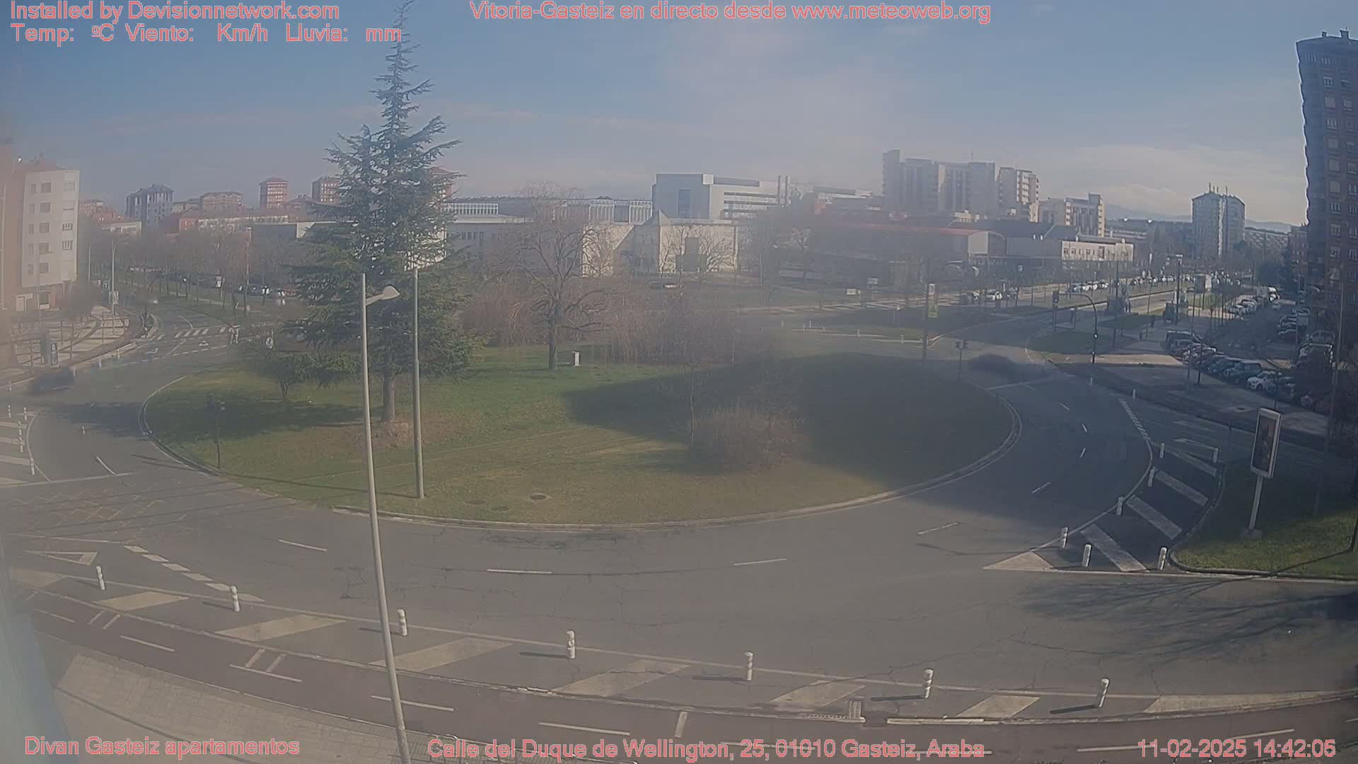 A sunny day reveals a traffic circle in a city, surrounded by buildings, green spaces, and parked cars.