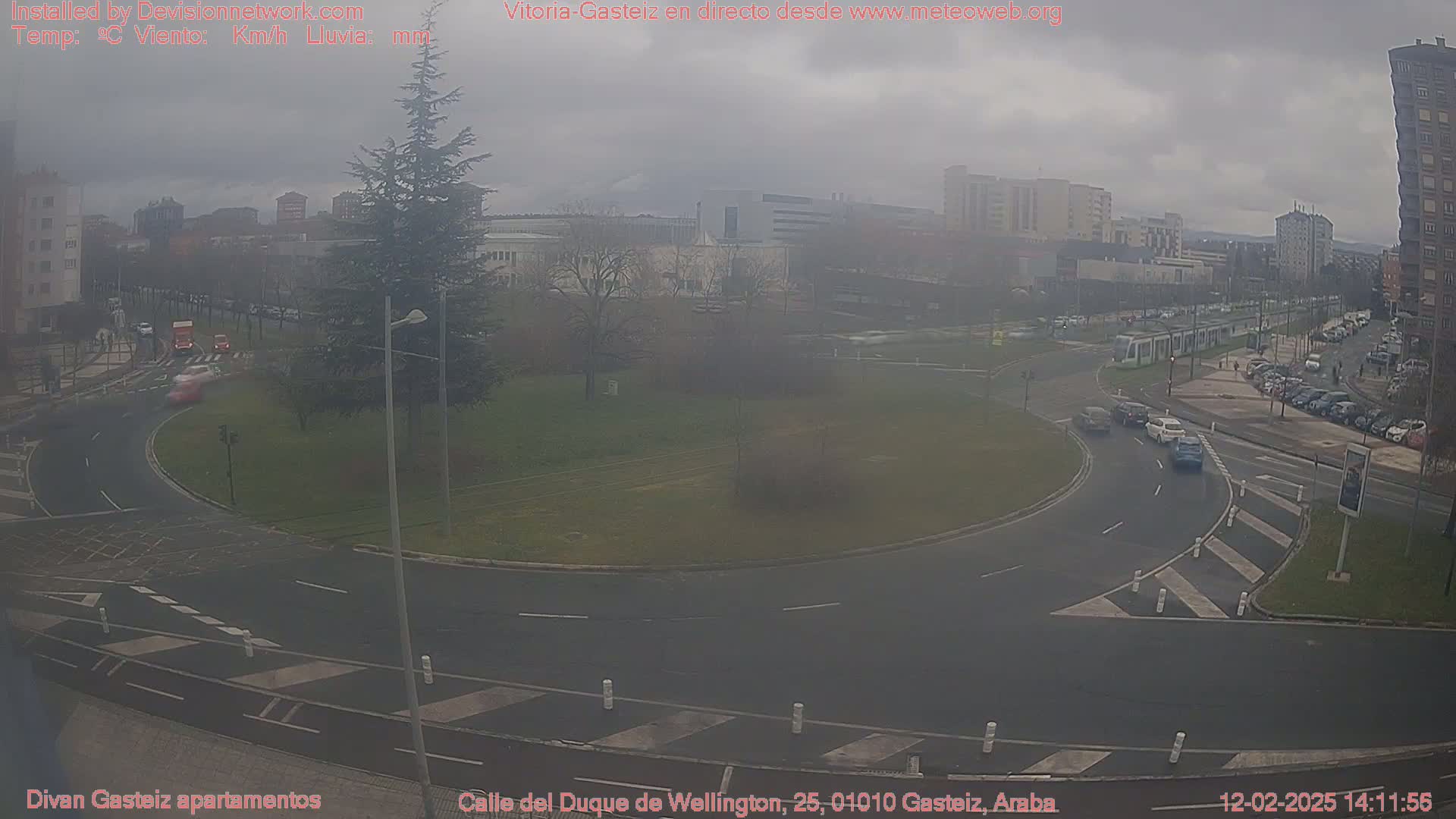 A hazy overcast day reveals a roundabout and surrounding city streets with several cars and a tram visible, all under a grey sky.