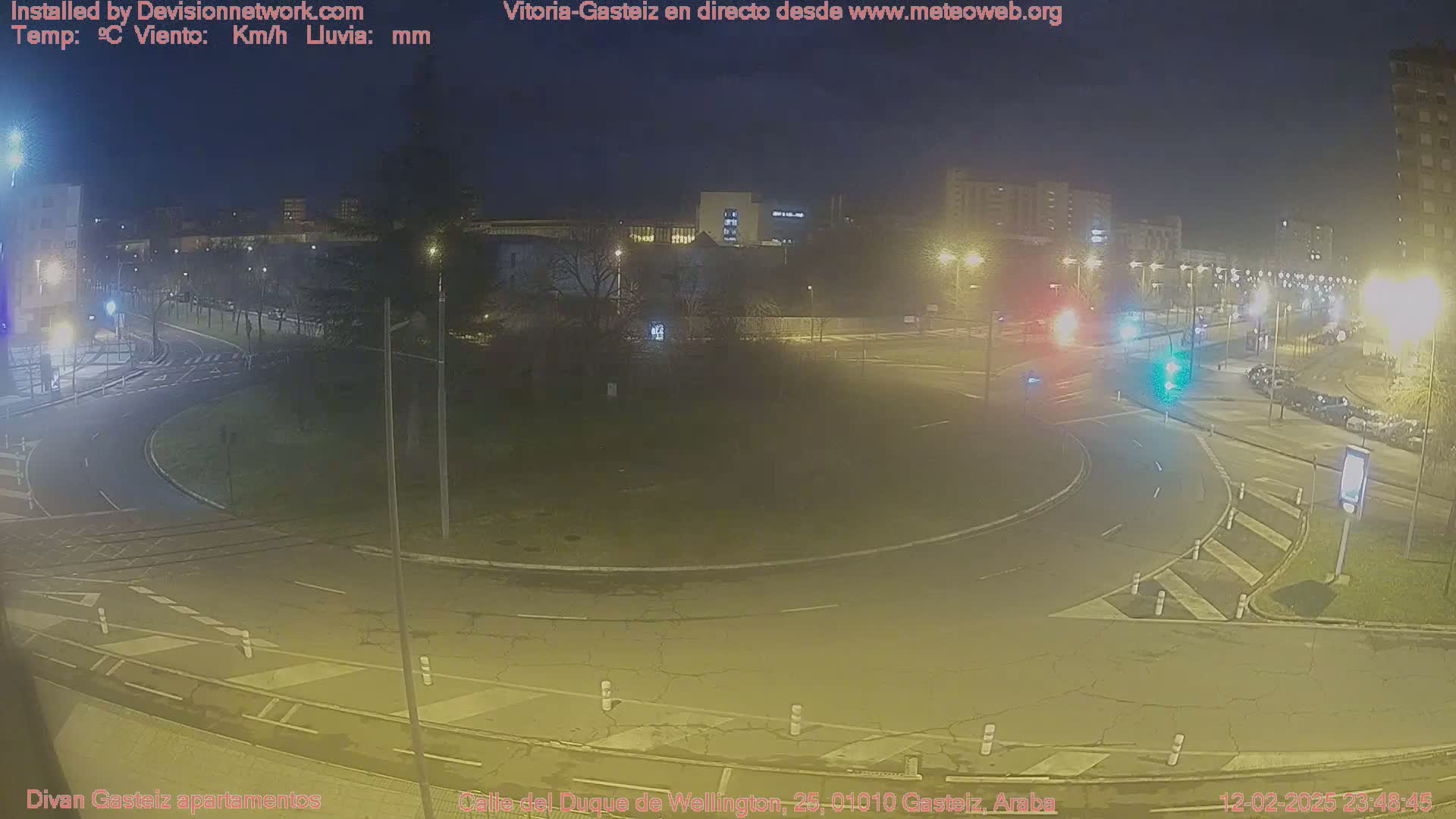 A nighttime, overhead view of a roundabout intersection with several cars and streetlights illuminating the scene.