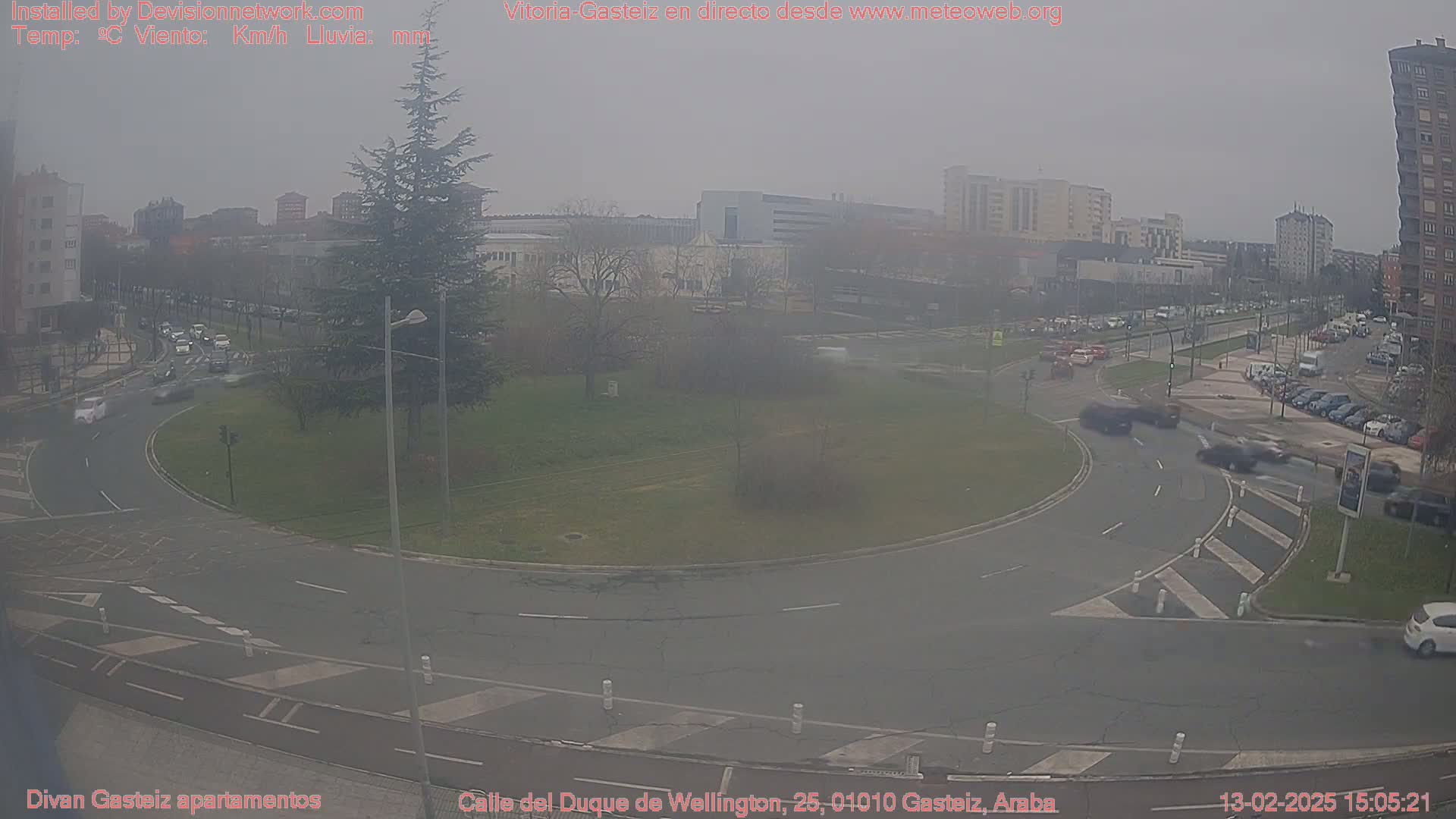 A hazy view of a roundabout in a city, with several cars visible, surrounded by buildings and trees.