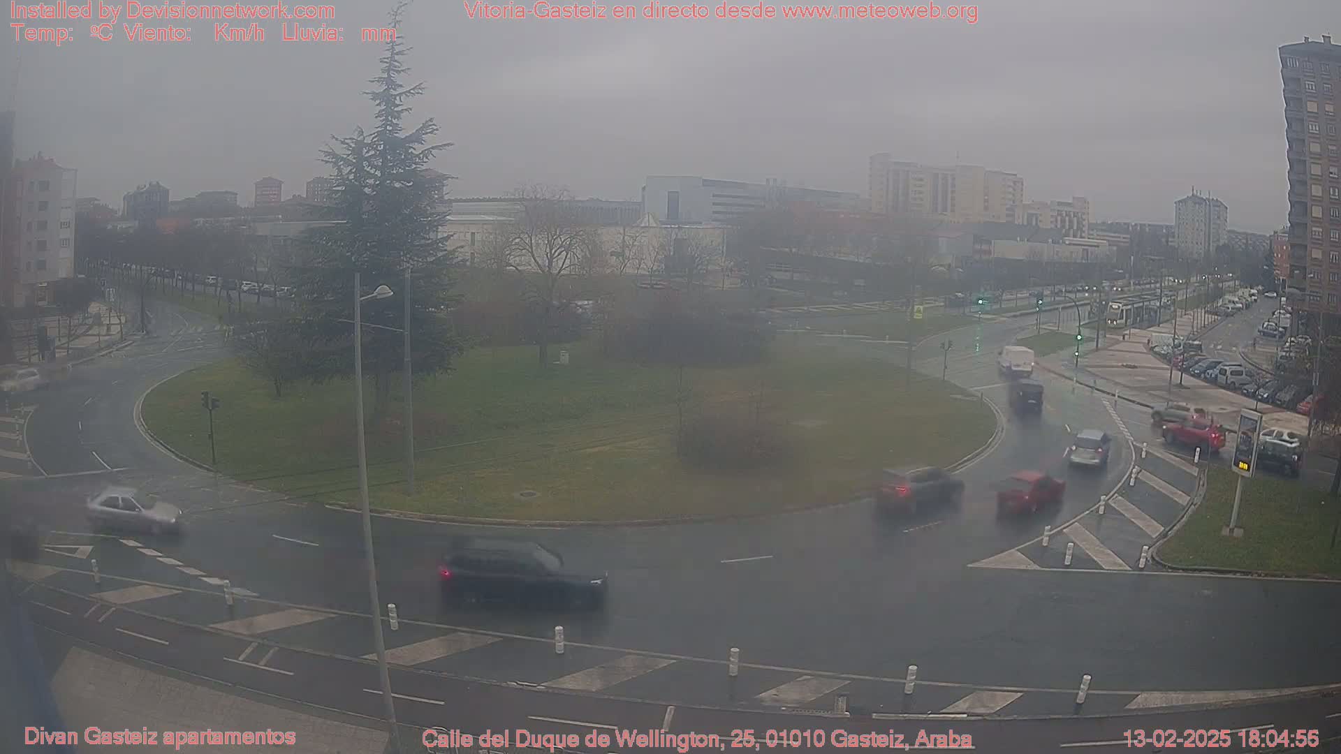A rainy, overcast day in a city roundabout, showing several cars navigating the intersection and buildings in the background.