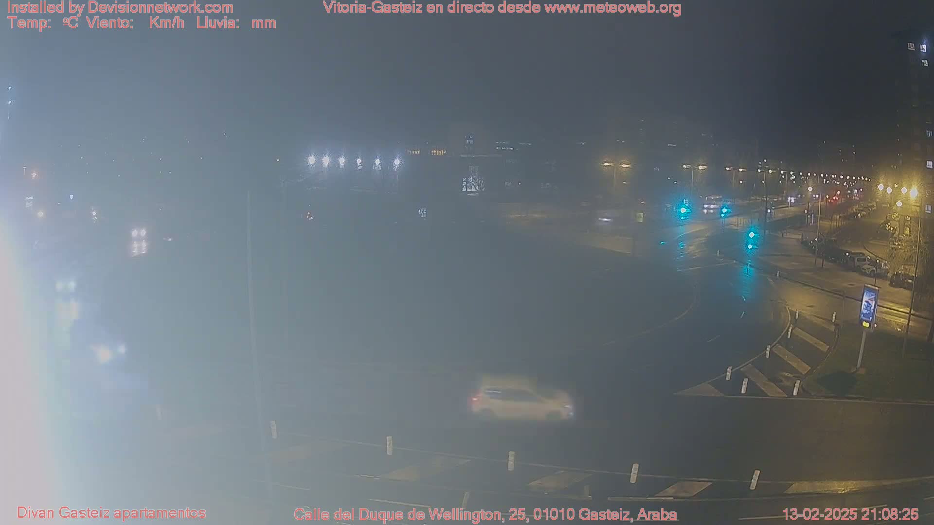 A nighttime, rainy view of a street intersection with several vehicles and streetlights, showing some fog or mist.