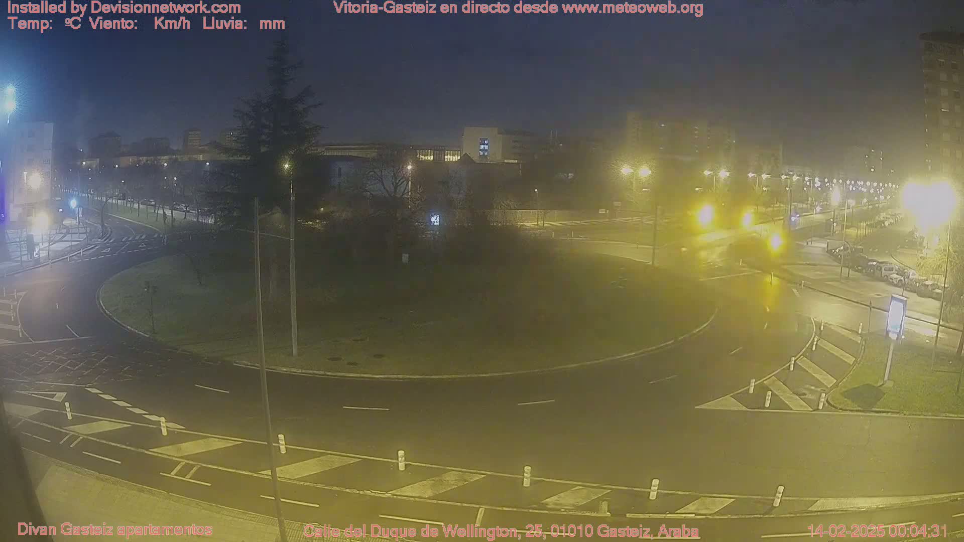 A nighttime view of a roundabout and surrounding streets with cars and streetlights, under wet conditions.