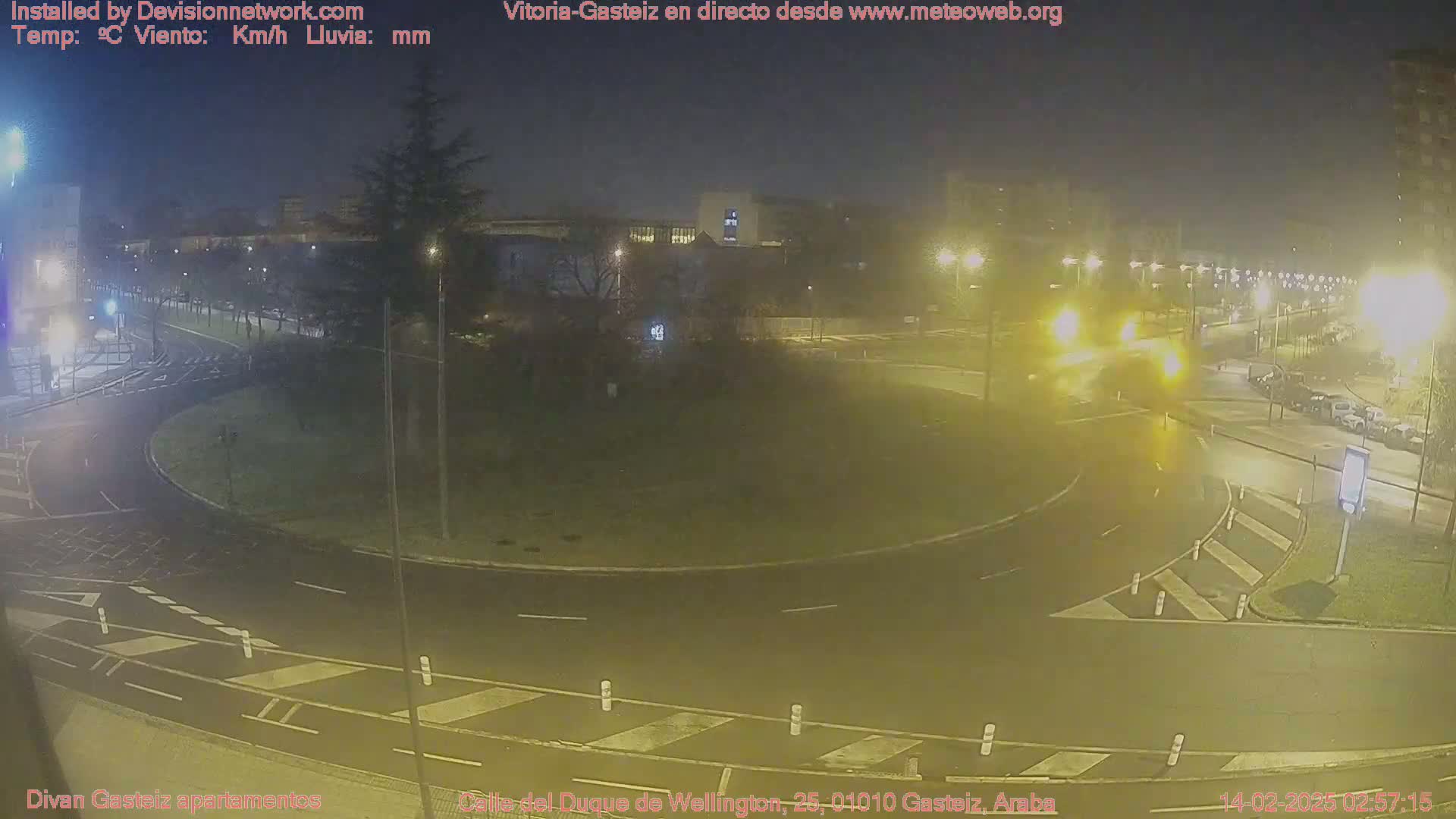 A nighttime view of a wet roundabout with a few cars and streetlights, under hazy conditions.