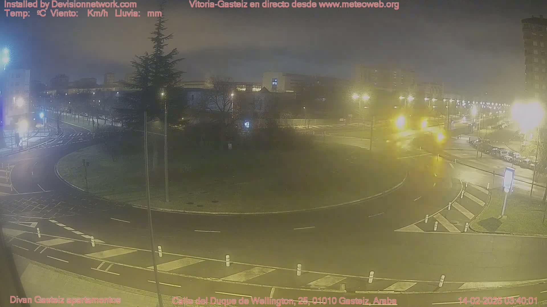 A nighttime view of a roundabout and surrounding roads with streetlights illuminating a slightly hazy and wet environment.