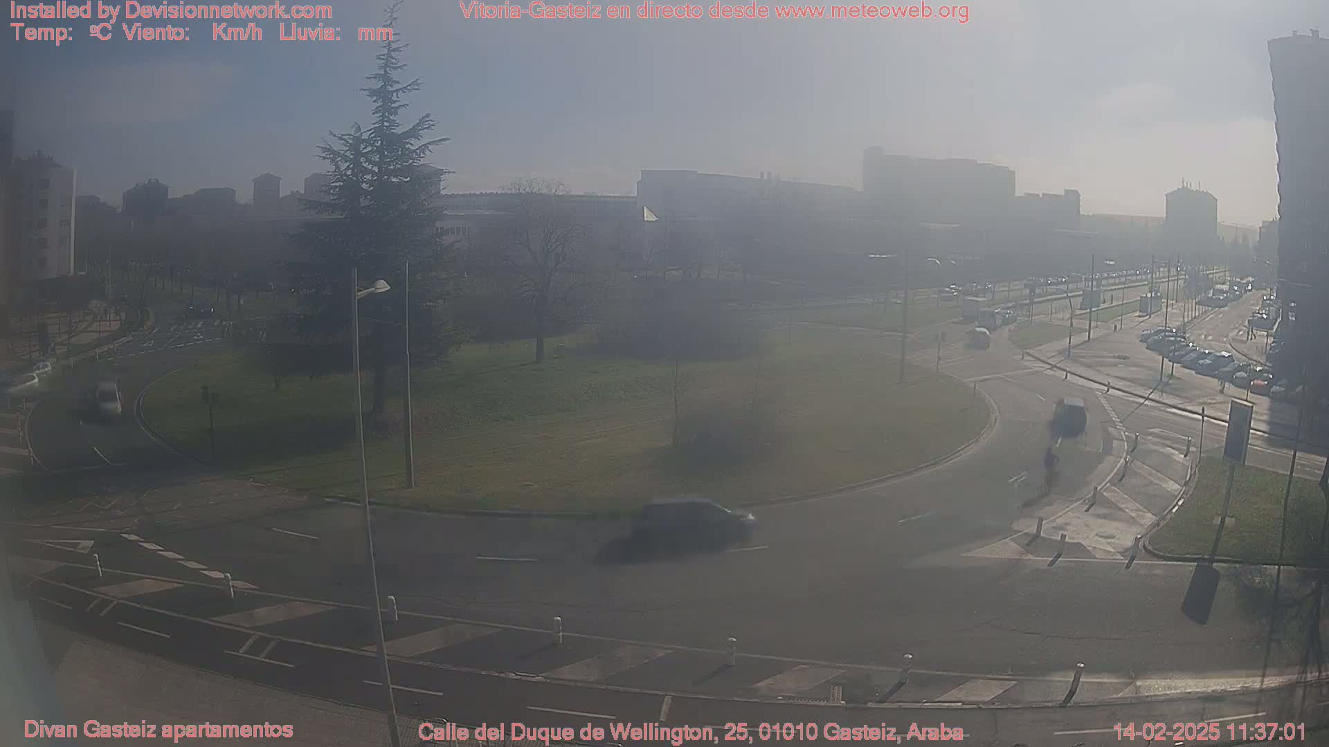 A hazy, sunny day shows a traffic circle and roads with several cars and pedestrians, surrounded by buildings and green spaces.