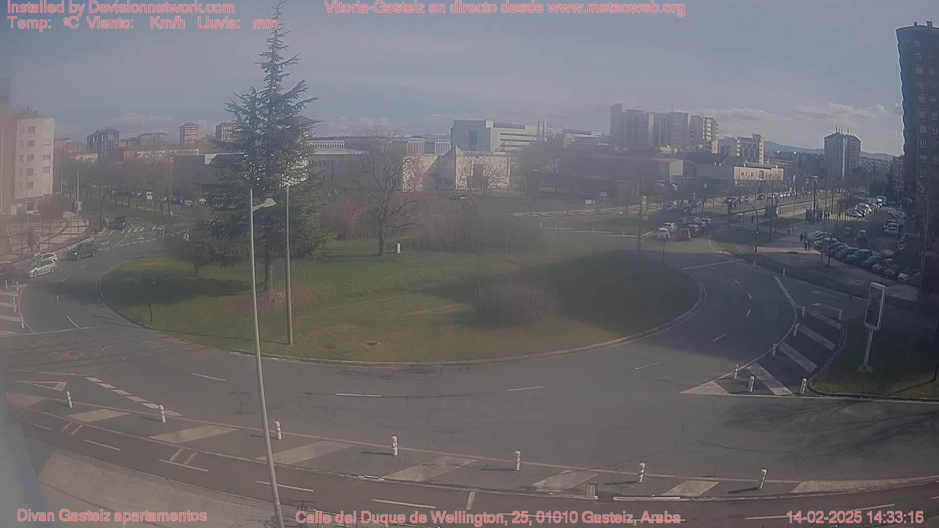 A sunny day reveals a roundabout in a city with several buildings and parked cars visible, along with some traffic moving through the roundabout.