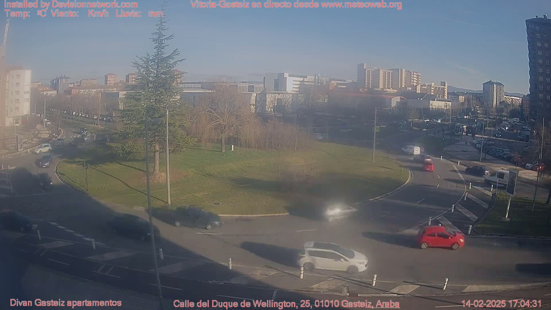 A sunny day shows a roundabout with several cars and a grassy area surrounded by buildings.