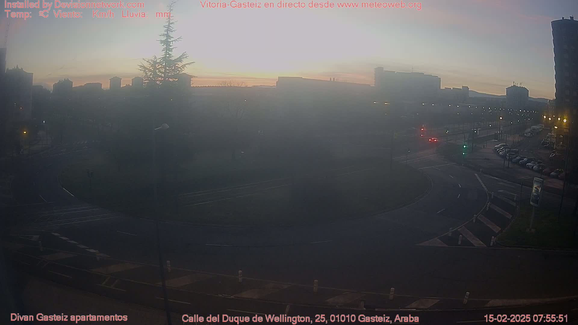 A hazy sunrise illuminates a roundabout and city streets with several cars visible; the atmosphere is foggy.