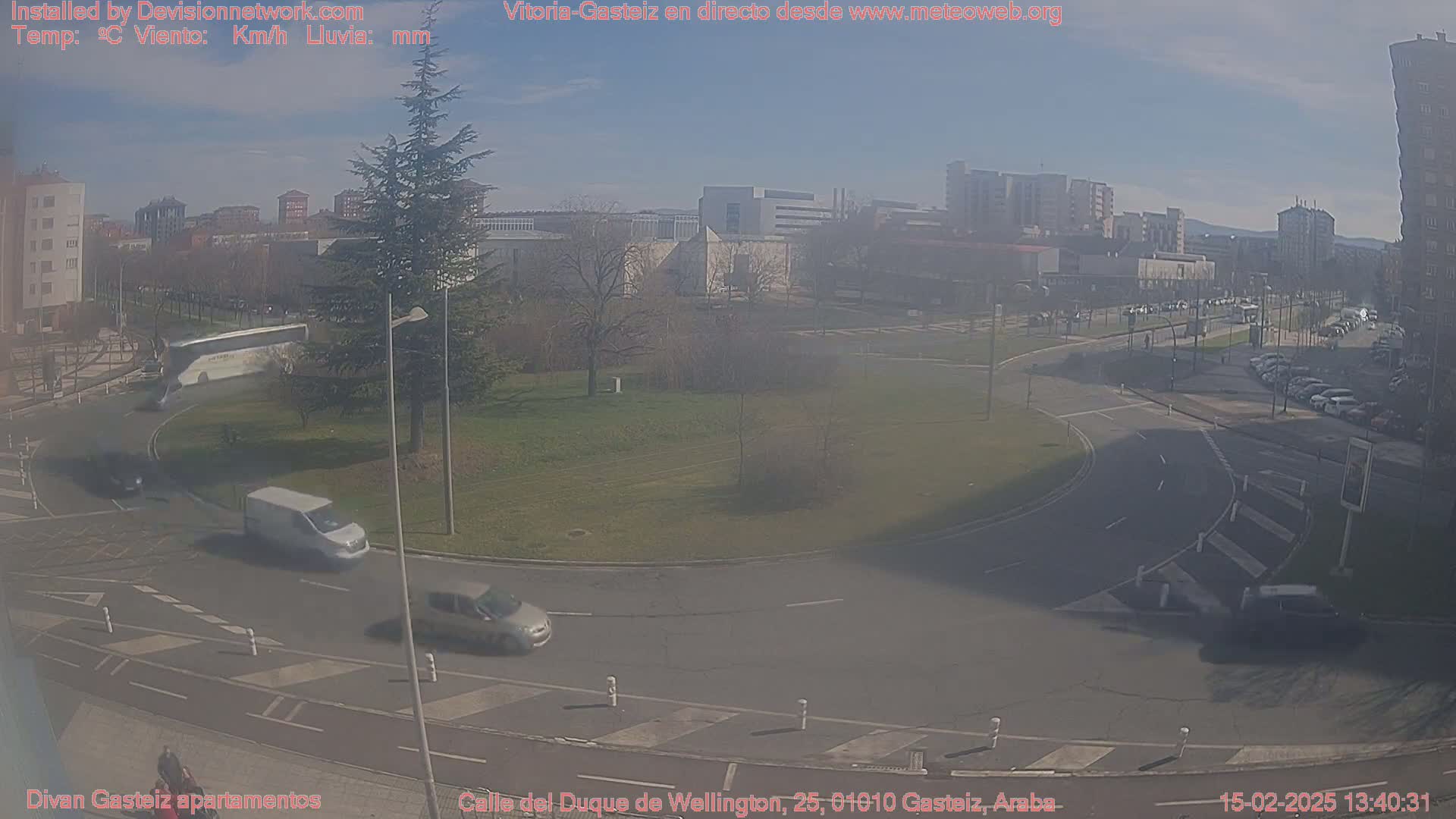 A sunny day shows a roundabout in a city with several cars driving around it, buildings in the background, and a large tree near the roundabout.