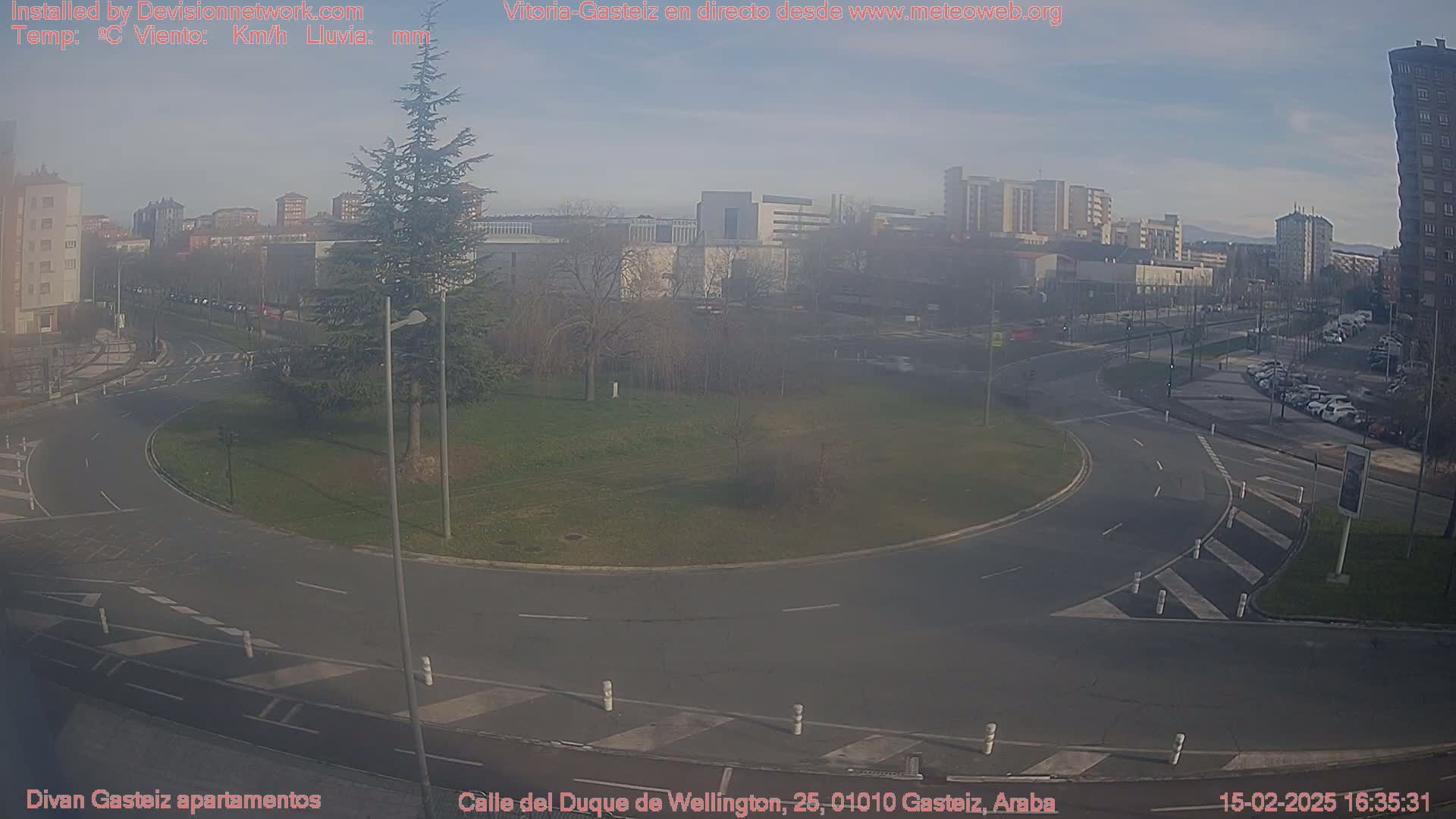 A hazy view of a roundabout and city streets, with buildings and trees visible in the background under a partly sunny sky.