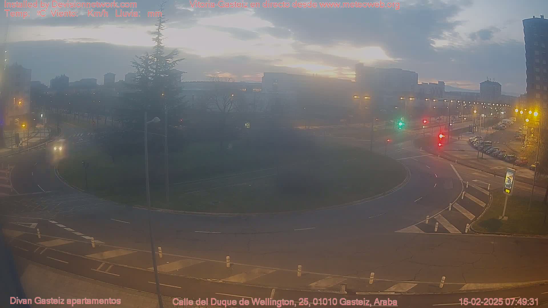 A hazy sunrise illuminates a roundabout and surrounding streets in a city, with several cars parked along the side of the road.