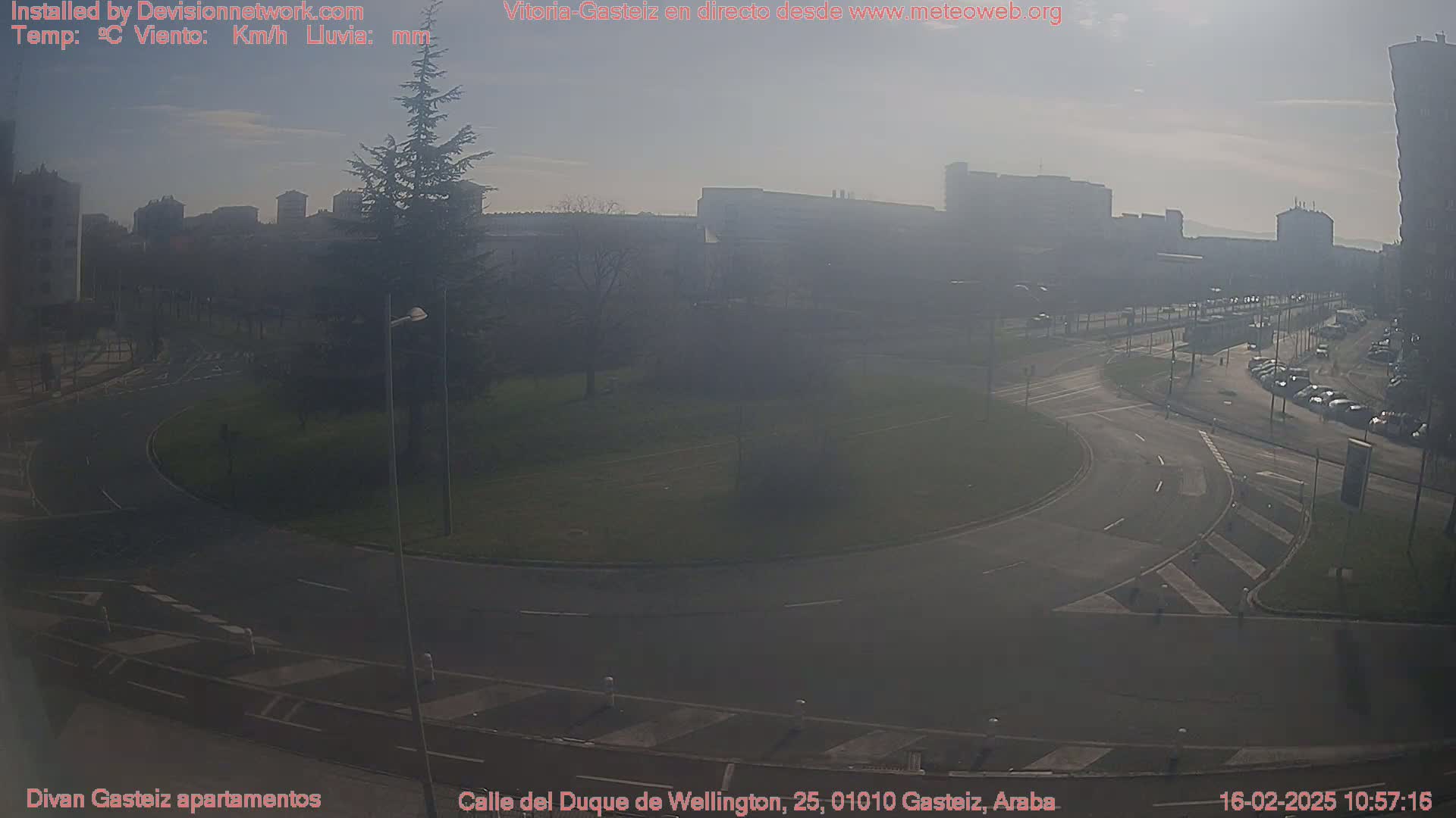 A hazy, sunny day reveals a roundabout and surrounding city streets, with several buildings and parked cars visible in the distance.