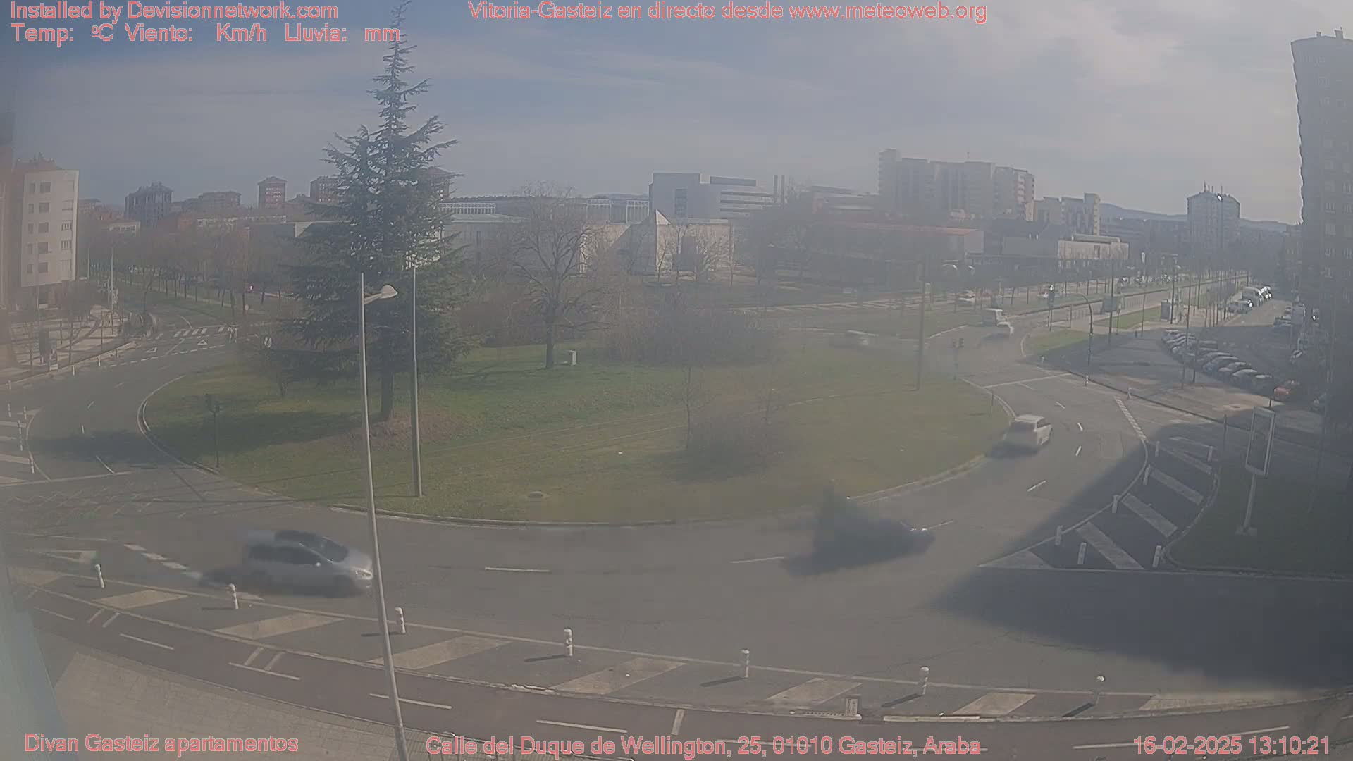 A hazy, sunny day reveals a roundabout and surrounding city streets, with several cars visible and buildings in the background.