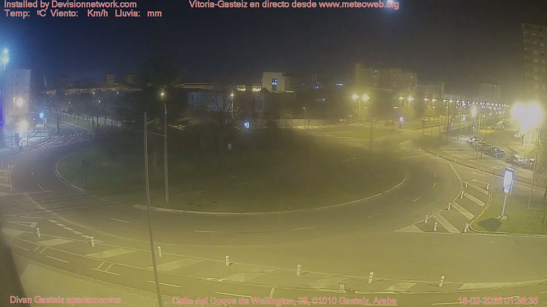 A nighttime, hazy view of a roundabout with cars parked nearby and buildings in the background.