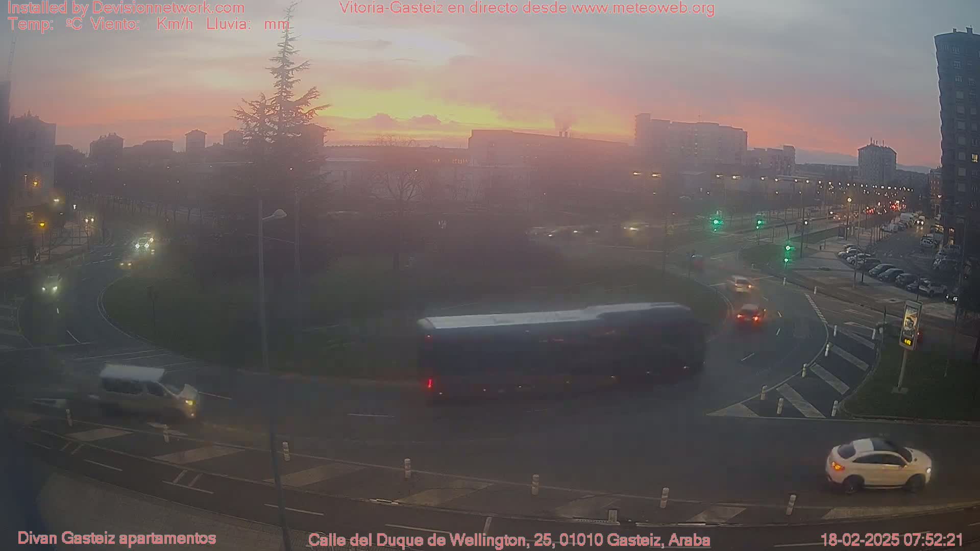 A city roundabout at sunrise, with several vehicles present, under a hazy, pinkish sky.