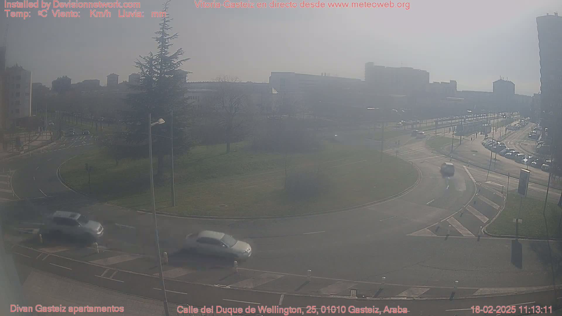 A hazy, sunny day reveals a roundabout with several cars, surrounded by buildings and green spaces.