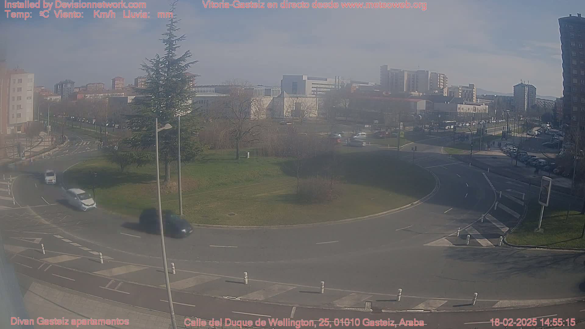 A sunny day reveals a roundabout surrounded by a grassy area, buildings, and vehicles moving through the intersection.