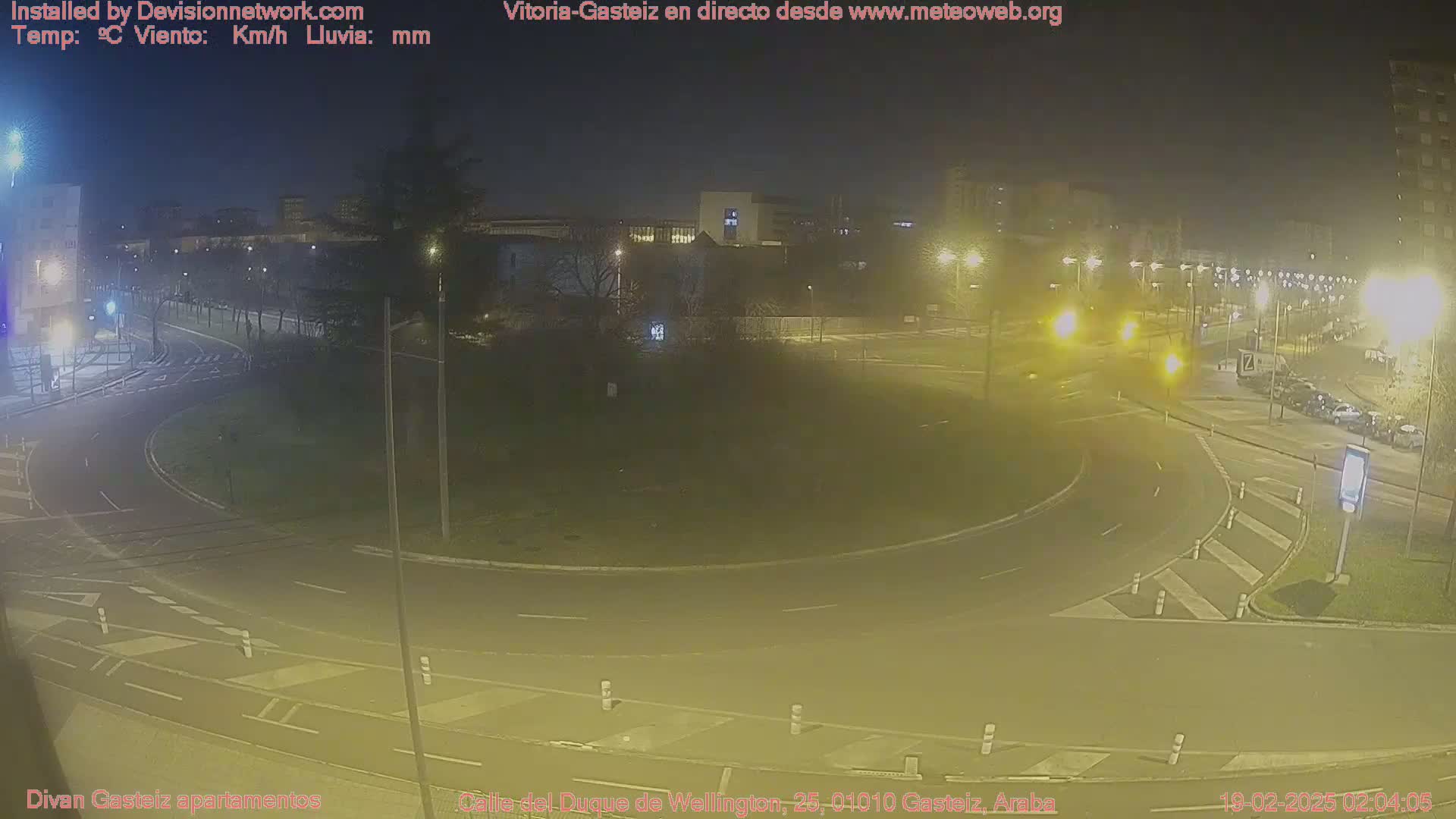 A nighttime view of a roundabout in a city with streetlights illuminating the road and surrounding buildings, under hazy conditions.