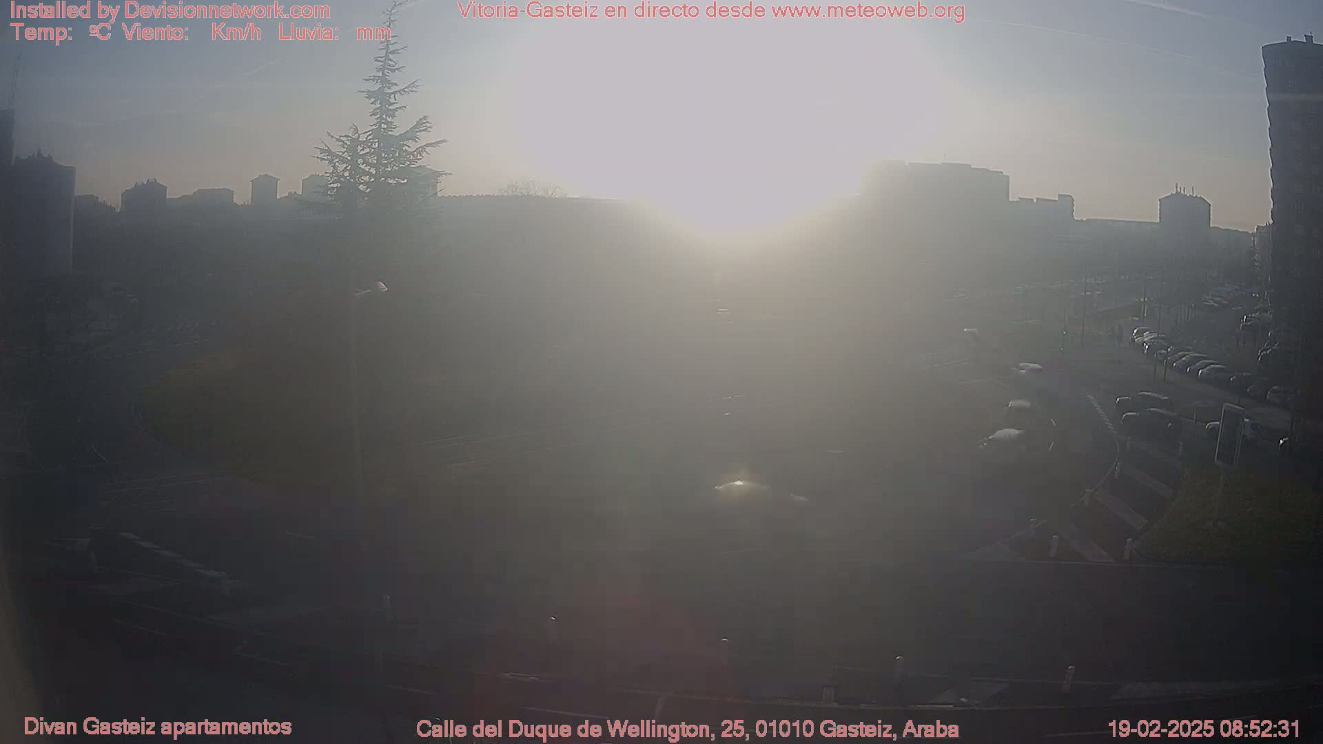 A hazy sunrise illuminates a city street with parked cars and buildings in the distance.