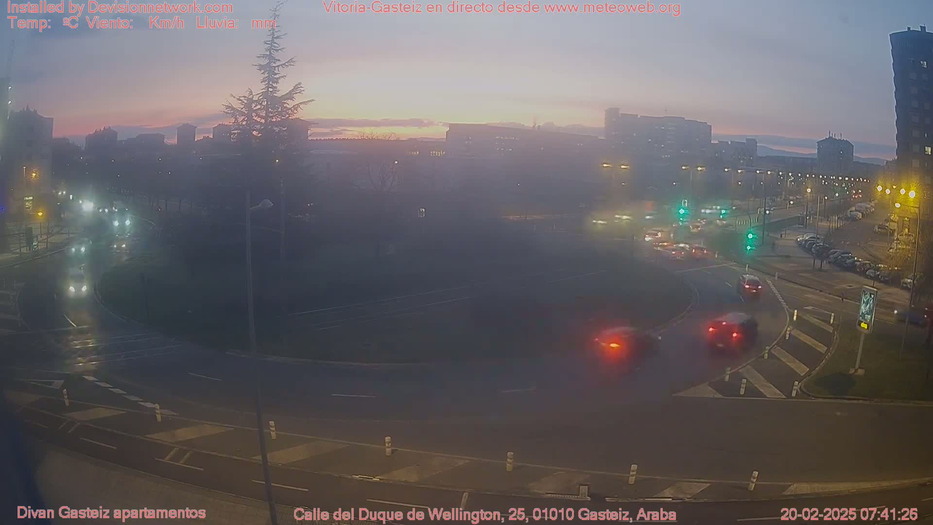 A twilight view of a roundabout with cars and buildings in the background, under hazy conditions.
