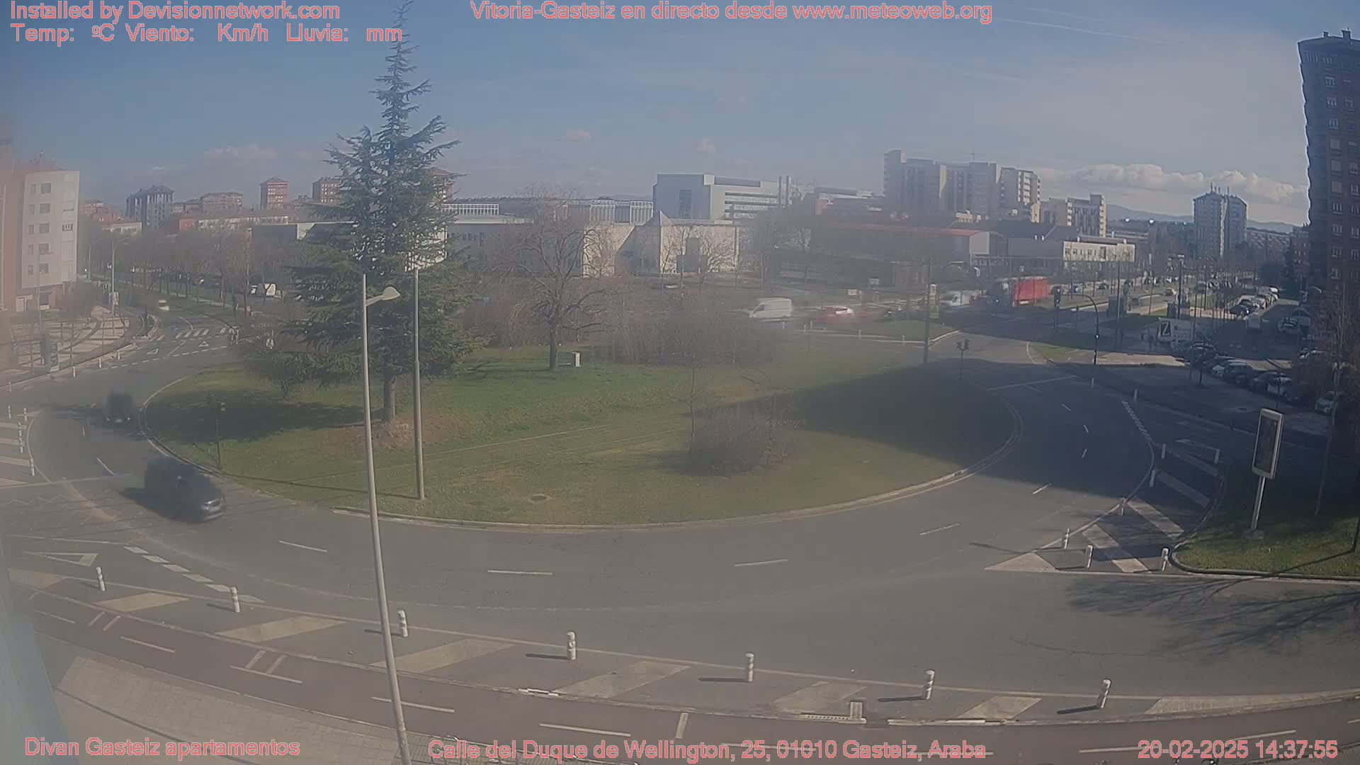 A sunny day reveals a traffic circle surrounded by a grassy area, buildings, and parked cars, with a few vehicles moving through the roundabout.
