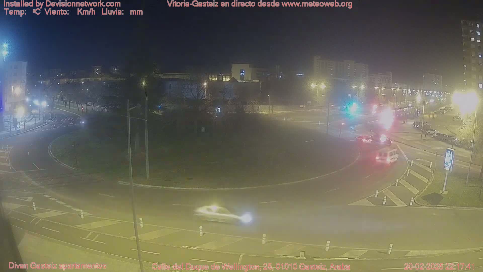 A nighttime view of a roundabout with several cars and buildings in the background under clear skies.