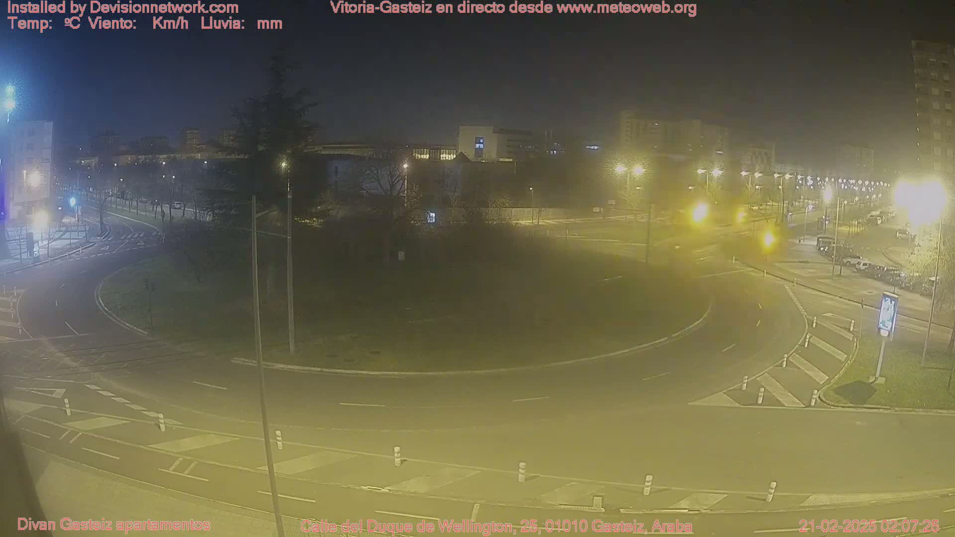 A nighttime view of a roundabout with sparse traffic and buildings in the background under hazy conditions.