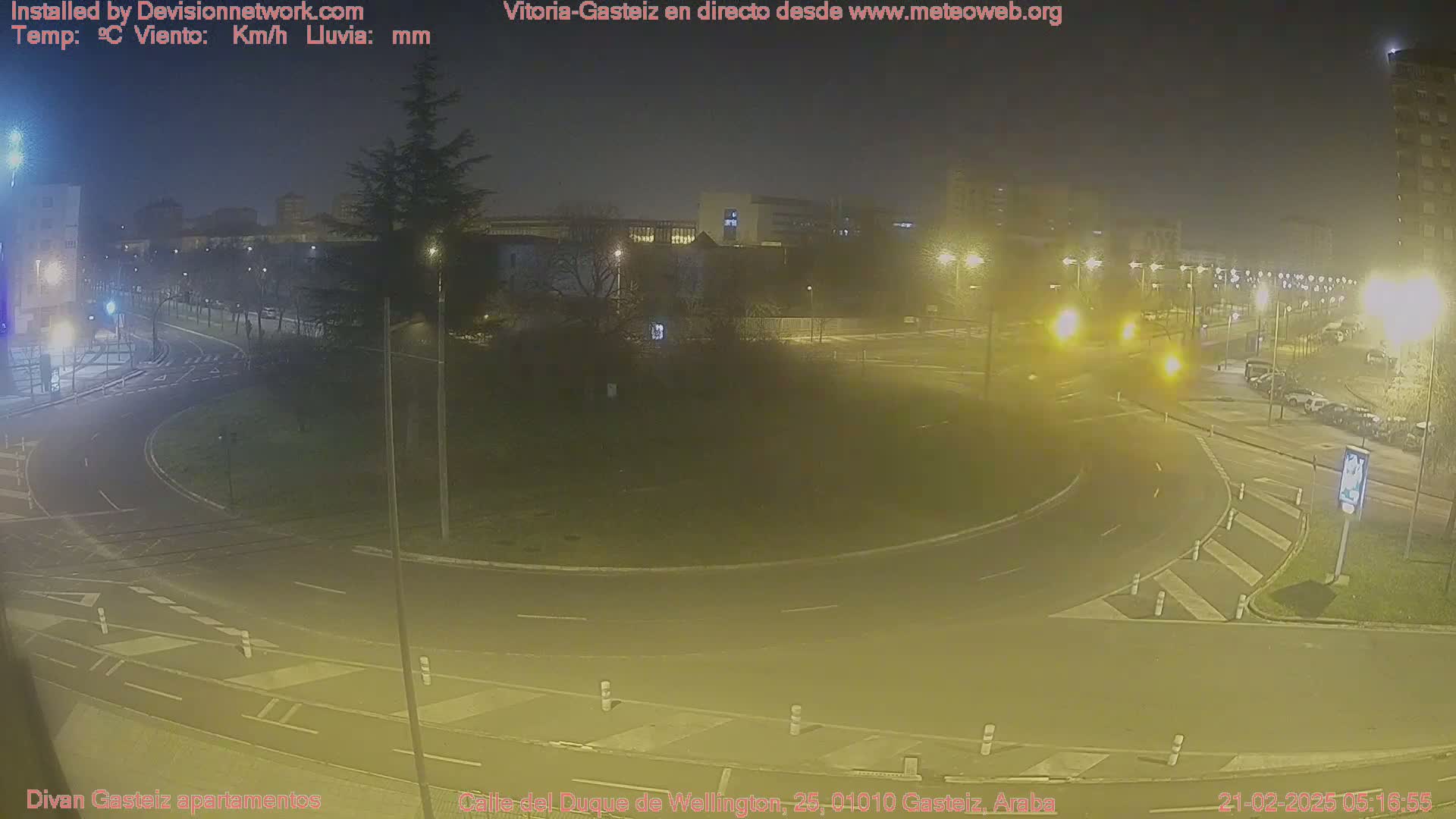 A nighttime, slightly hazy view of a roundabout in a city, with streetlights illuminating the road and surrounding buildings.