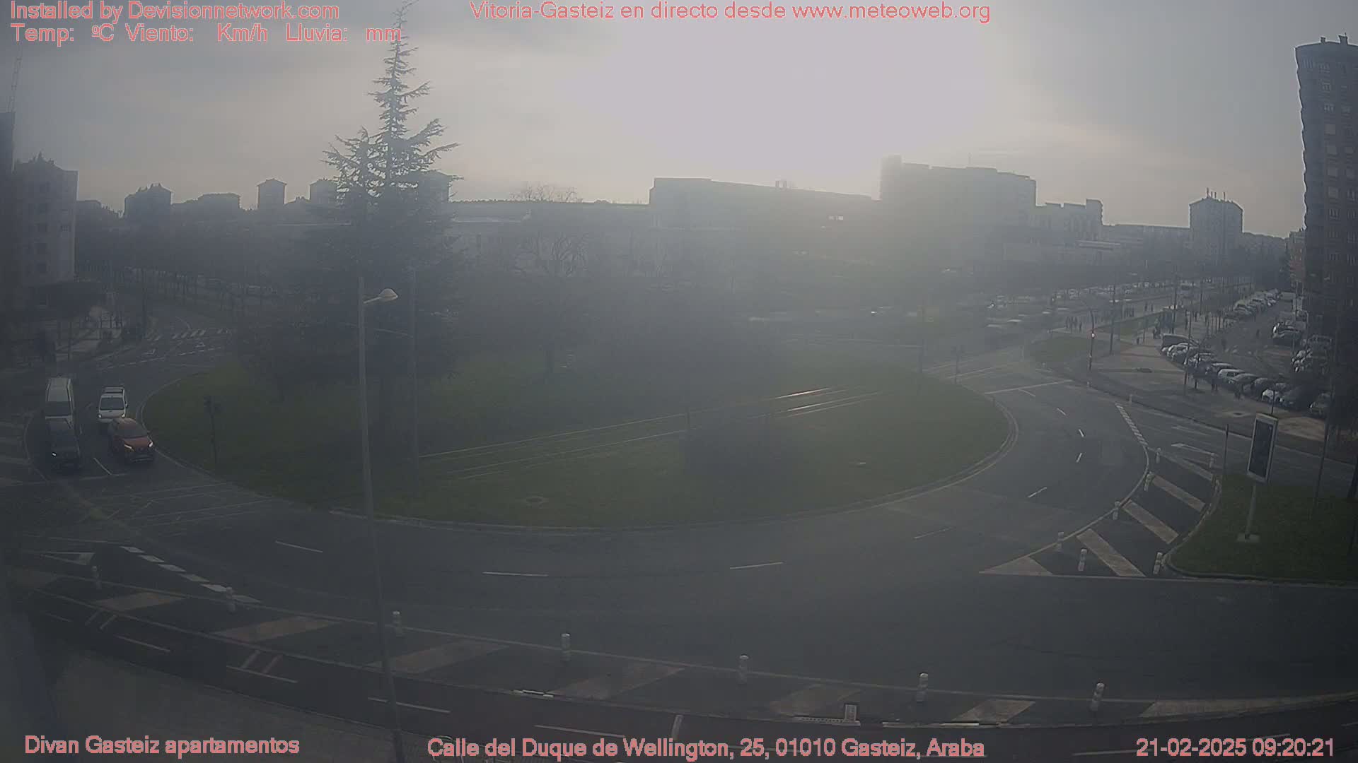 A hazy view of a city roundabout with several cars, surrounded by buildings and a tree, on a partly sunny day.