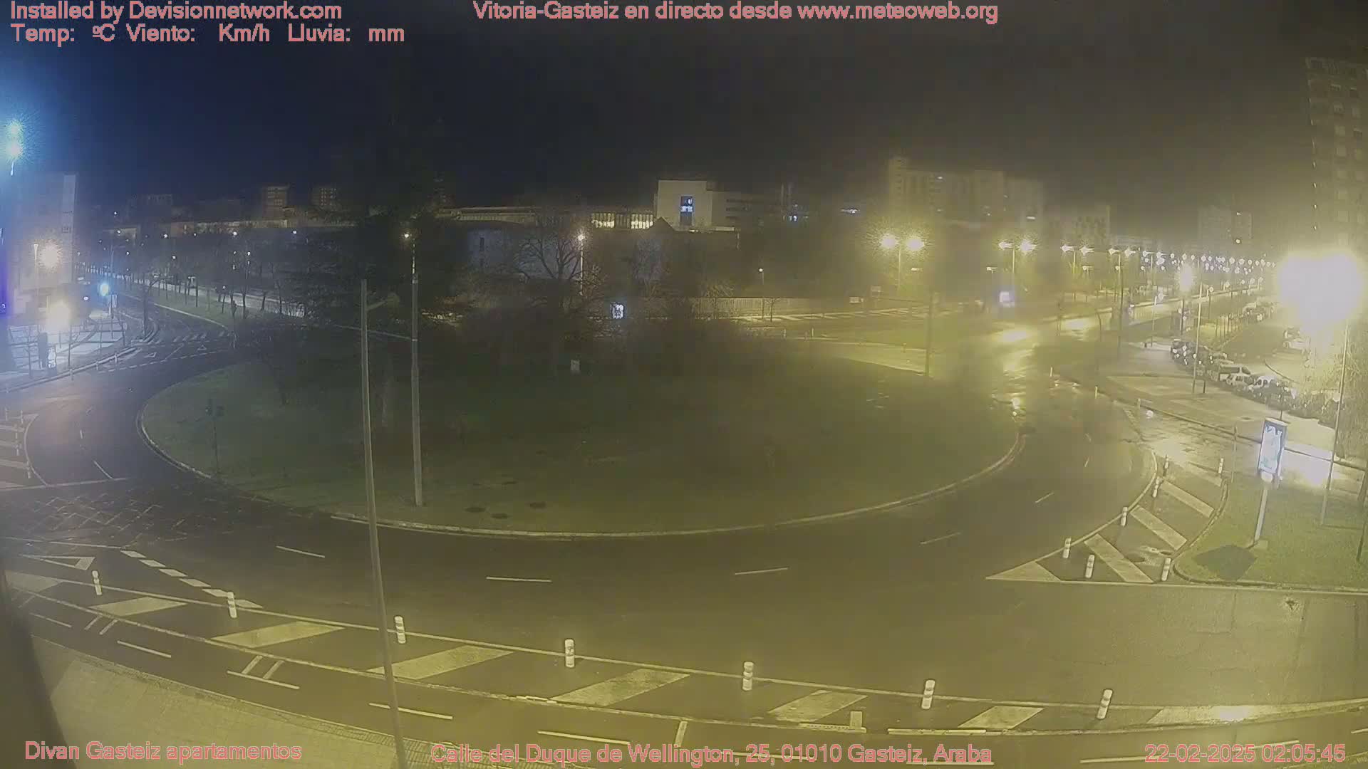 A nighttime view of a wet roundabout in a city, with streetlights illuminating the surrounding area and parked cars visible.