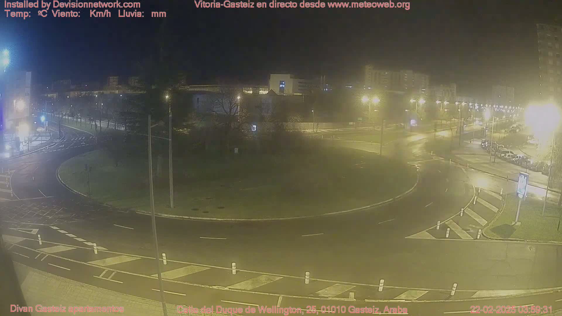 A nighttime view of a roundabout and surrounding streets with several cars visible, under wet conditions.