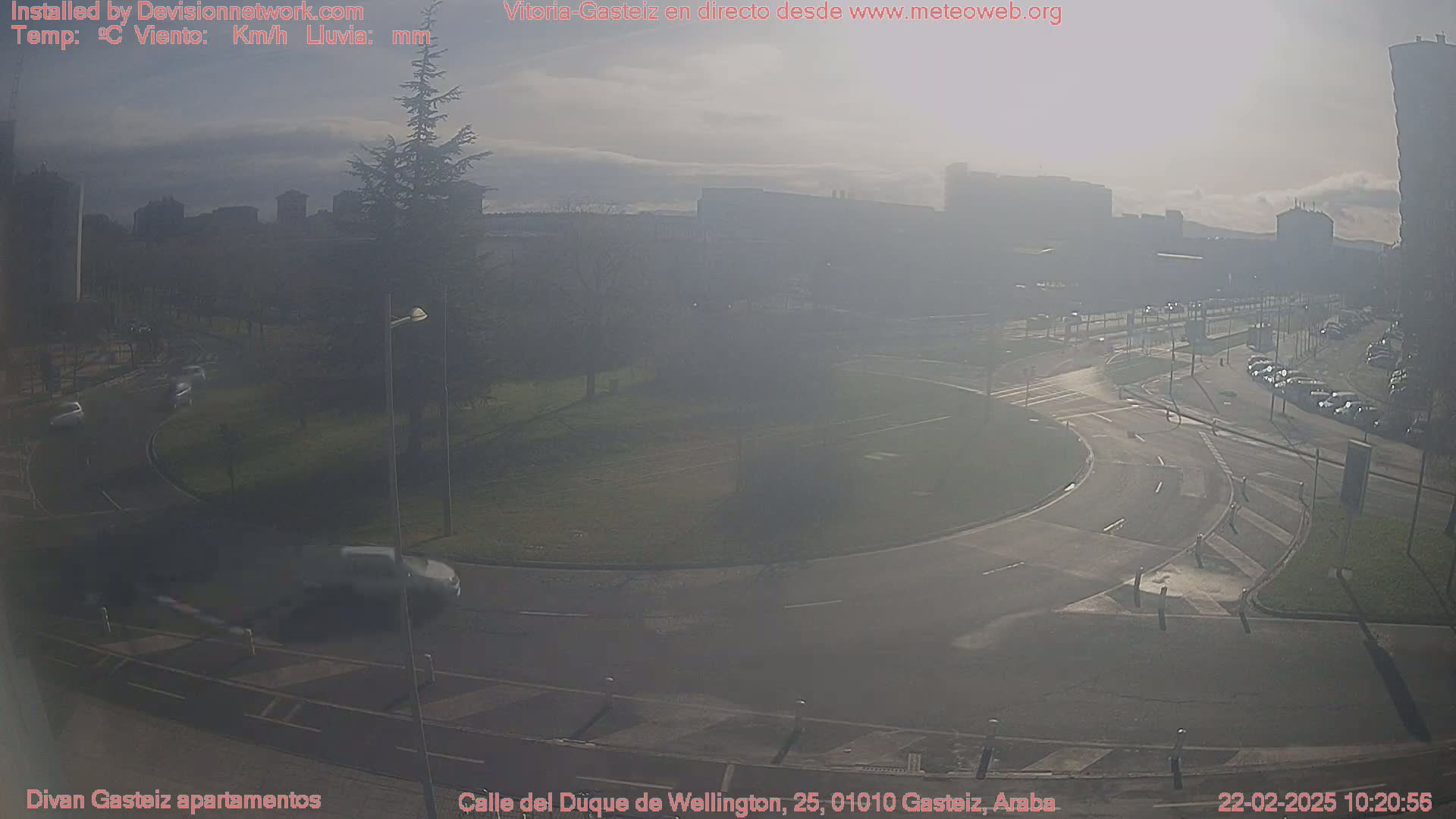 A hazy, sunny day reveals a roundabout and roads with sparse traffic, surrounded by buildings and a small park.