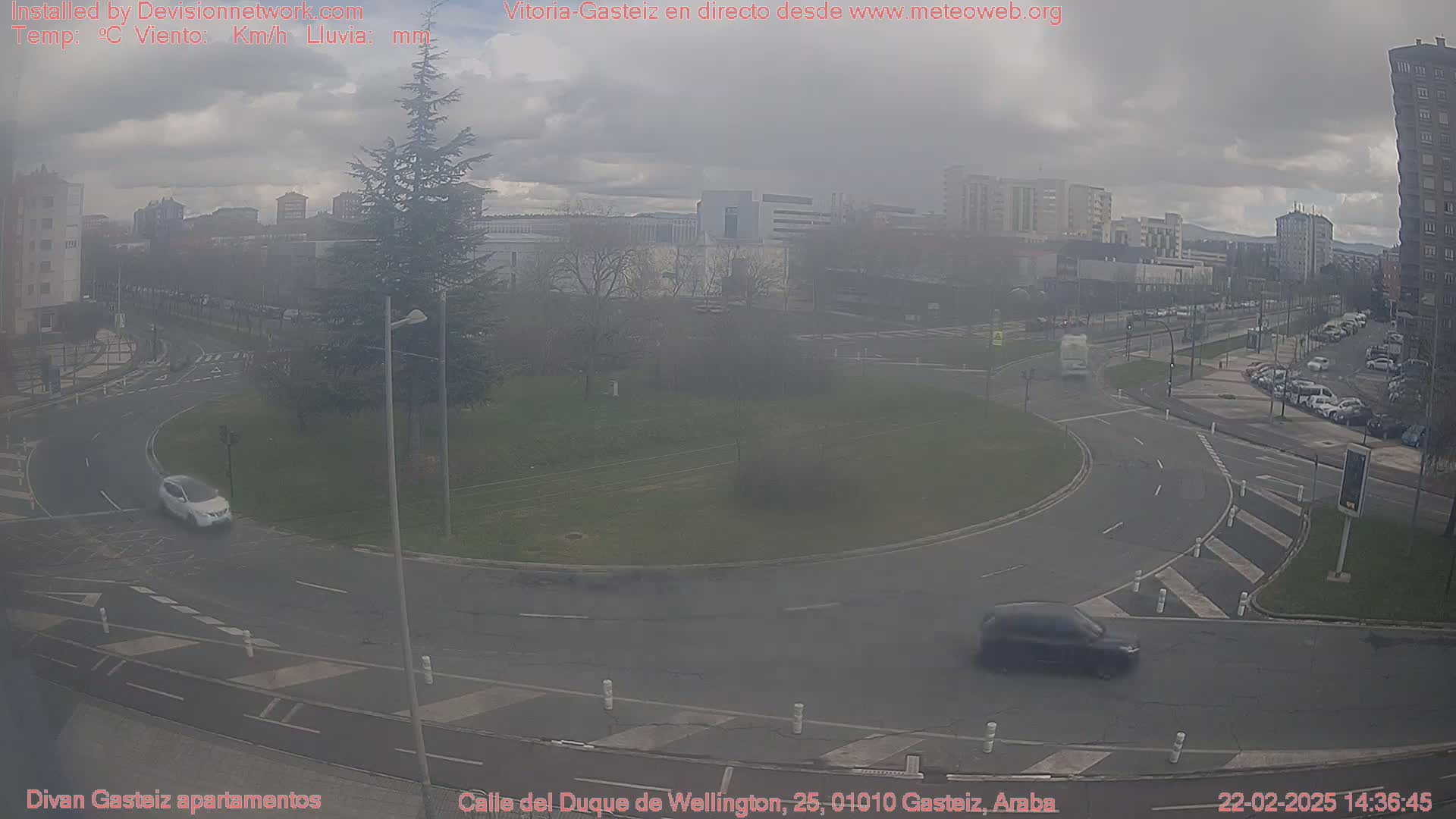 A roundabout in an urban area on an overcast day, with several cars visible on the roads and in a nearby parking lot.