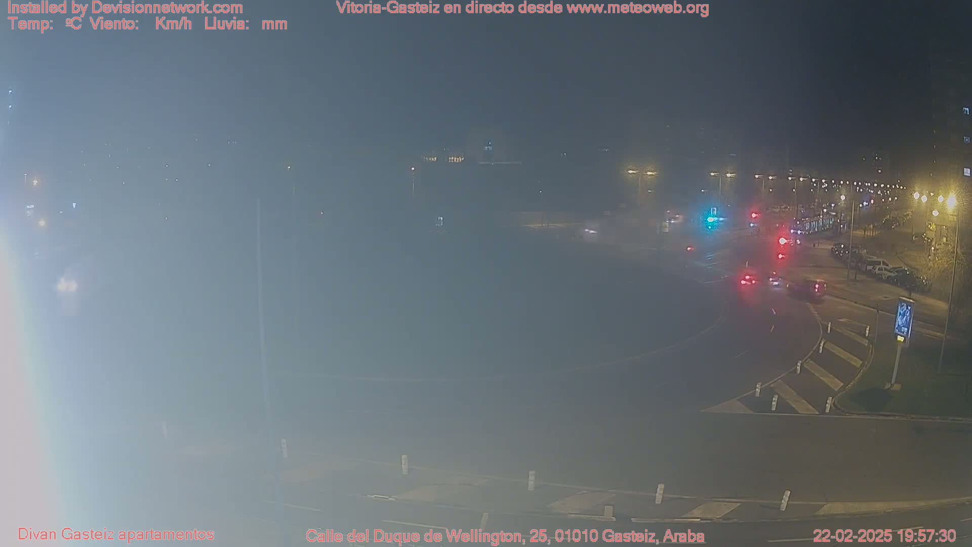 A nighttime, hazy view of a roundabout with several cars and streetlights.