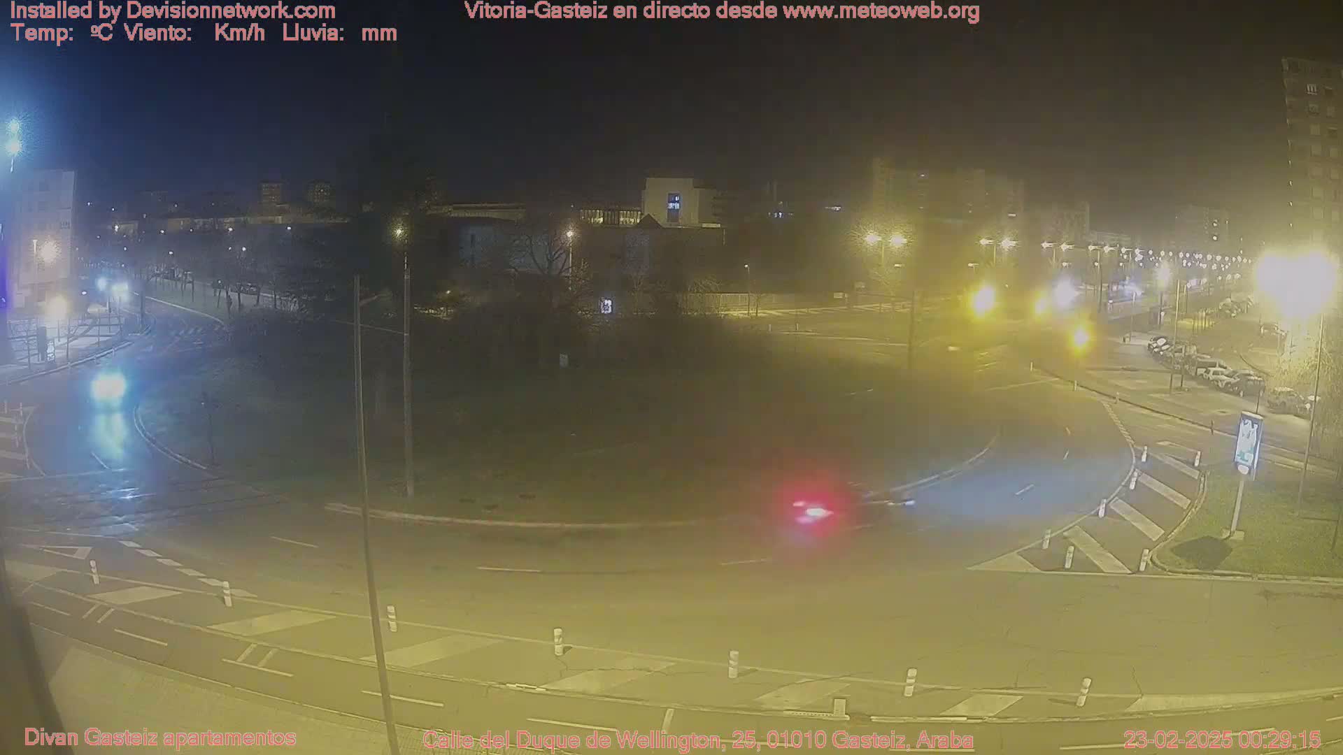 A wet roundabout at night with cars driving through it and buildings visible in the background under dim streetlights.