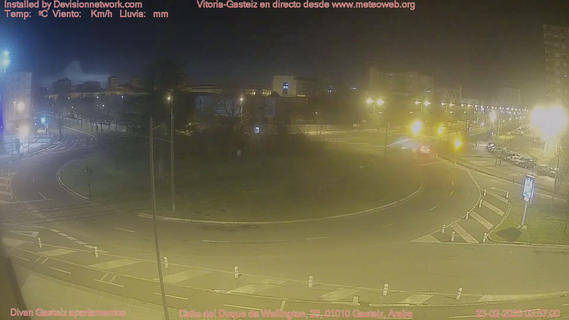 A nighttime view of a roundabout in a city, illuminated by streetlights, with some parked cars visible.