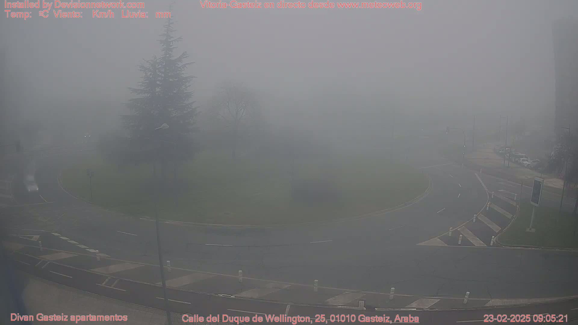 A foggy view of a roundabout and surrounding roads, with a few cars visible and trees partially obscured by the dense fog.