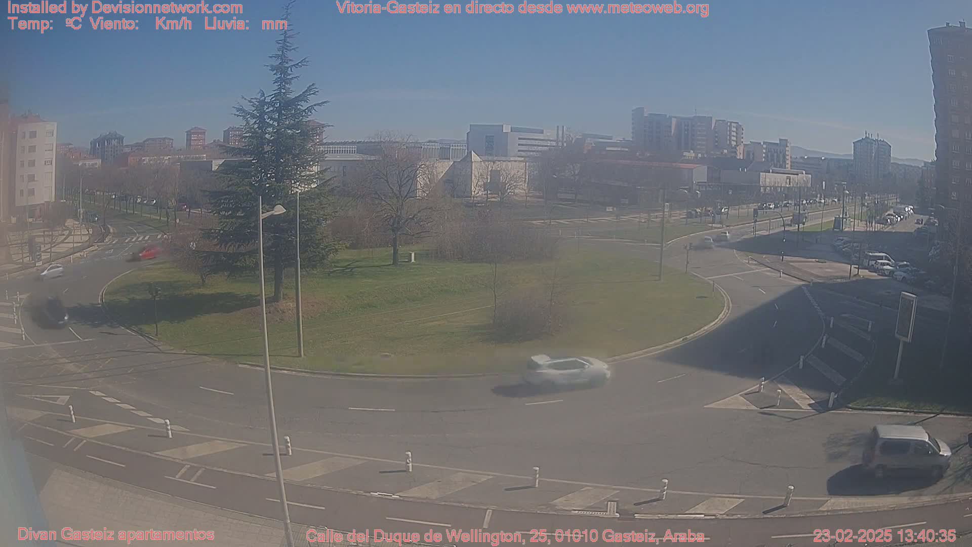 A sunny day overlooking a roundabout with several cars, surrounded by buildings and a grassy area with trees.
