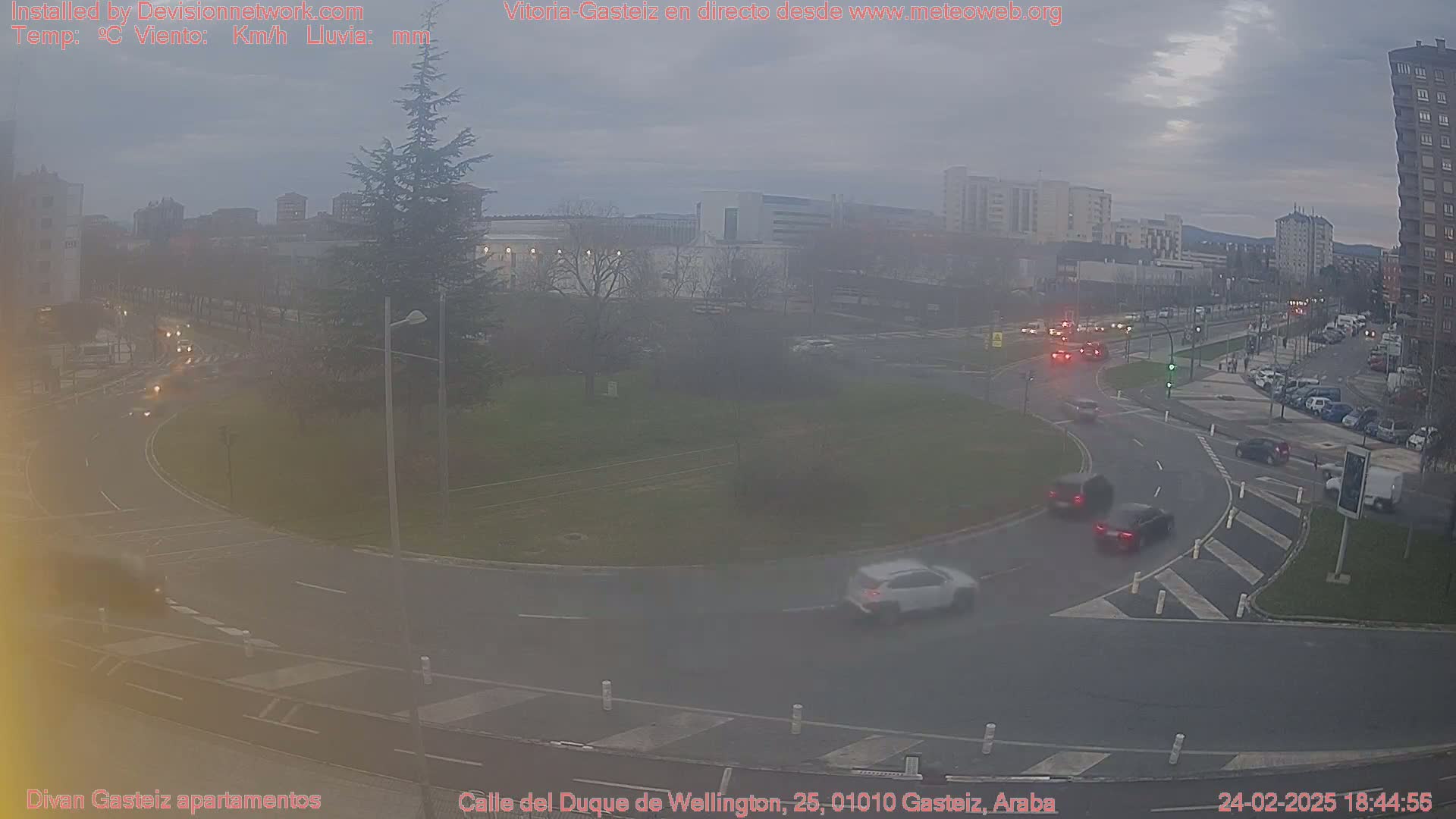 A hazy, overcast day reveals a roundabout with several cars navigating it, surrounded by buildings and trees.