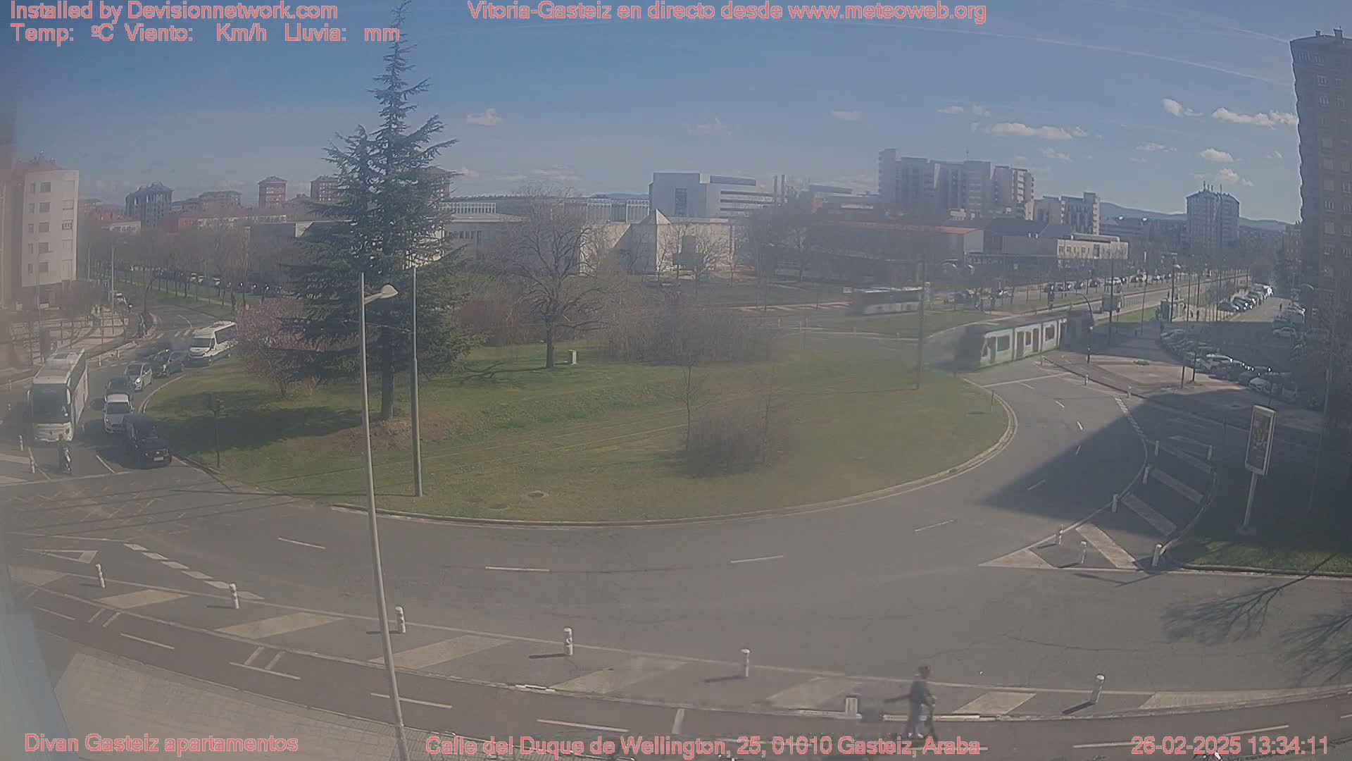 A sunny day reveals a street intersection with a tram, several vehicles, and pedestrians, surrounded by buildings and green spaces.