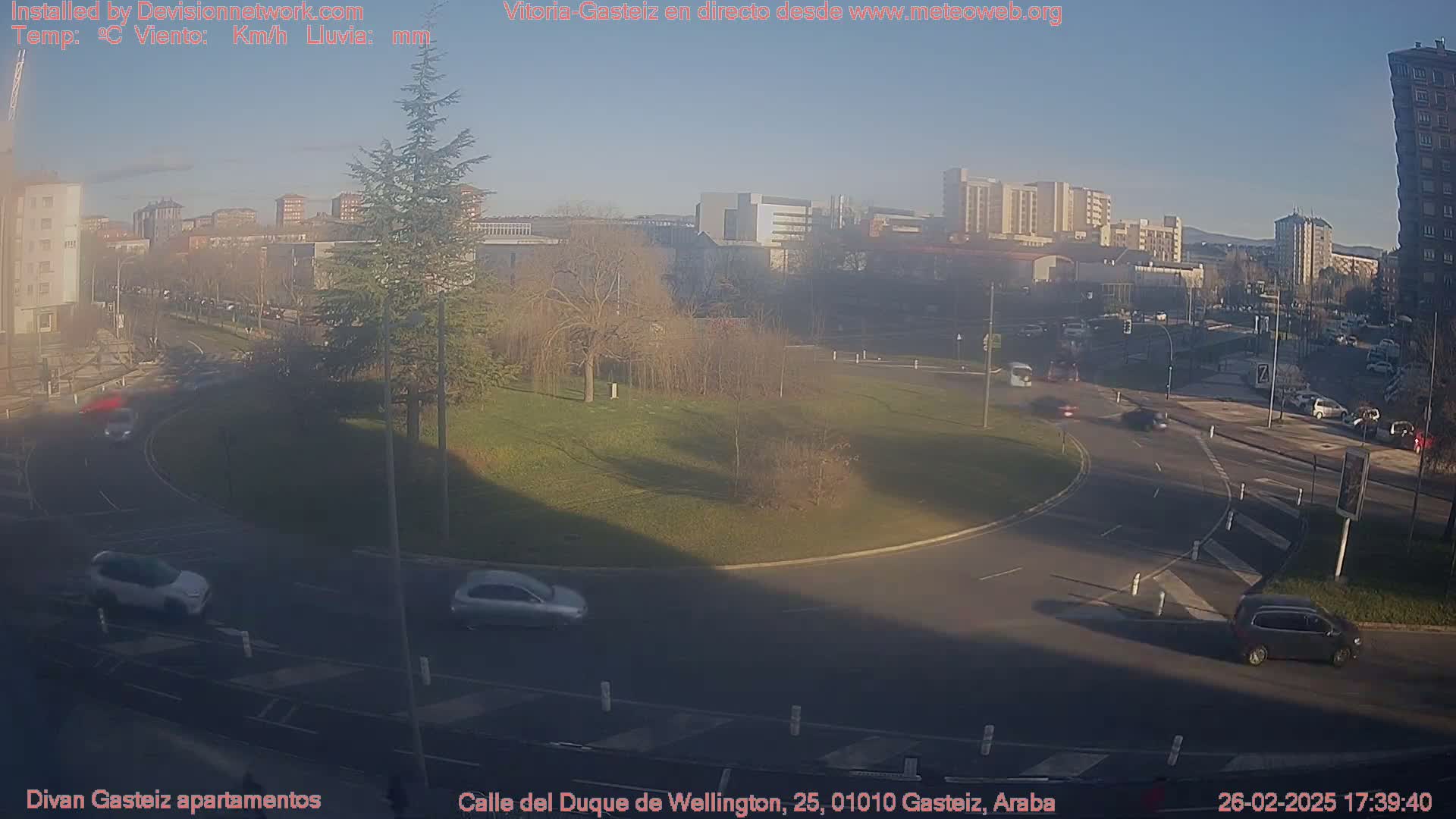 A mostly sunny view of a roundabout with several cars driving on the roads and a grassy area with trees in the center, surrounded by buildings in a city.