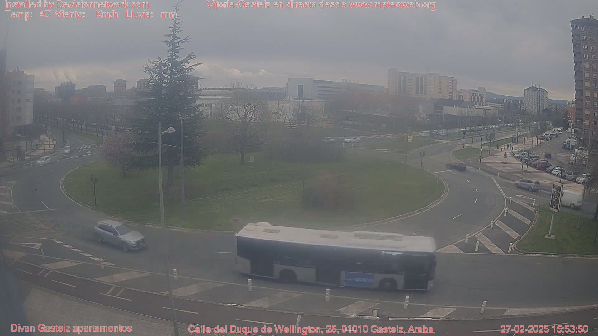 A city street scene on an overcast day shows a roundabout with cars and a bus, near buildings and trees.