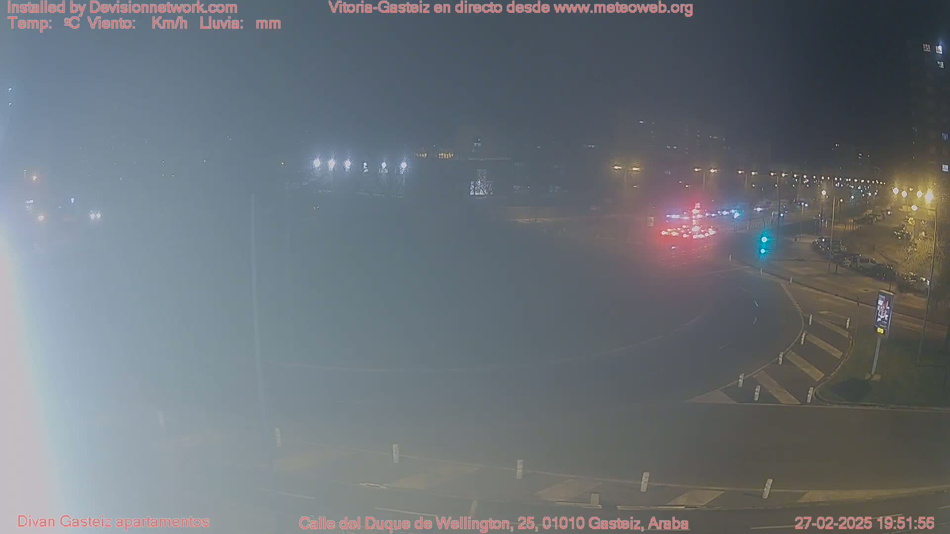 A nighttime, foggy view of a traffic circle with several cars and streetlights visible.