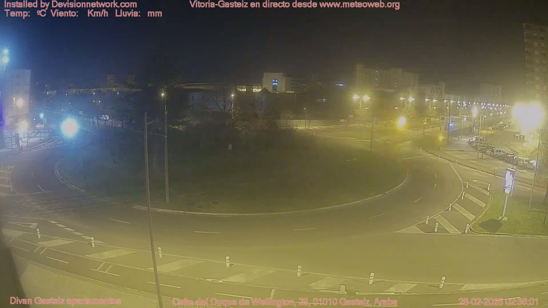 A nighttime view of a roundabout in a city with some cars, streetlights, and buildings in the background under hazy conditions.