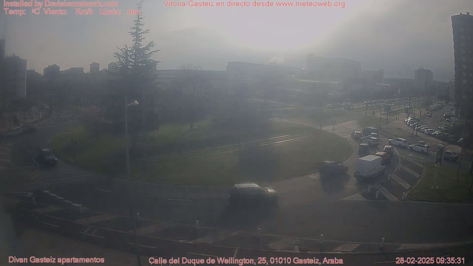 A hazy, sunlit roundabout and surrounding streets show several cars and a van moving through a somewhat busy intersection.