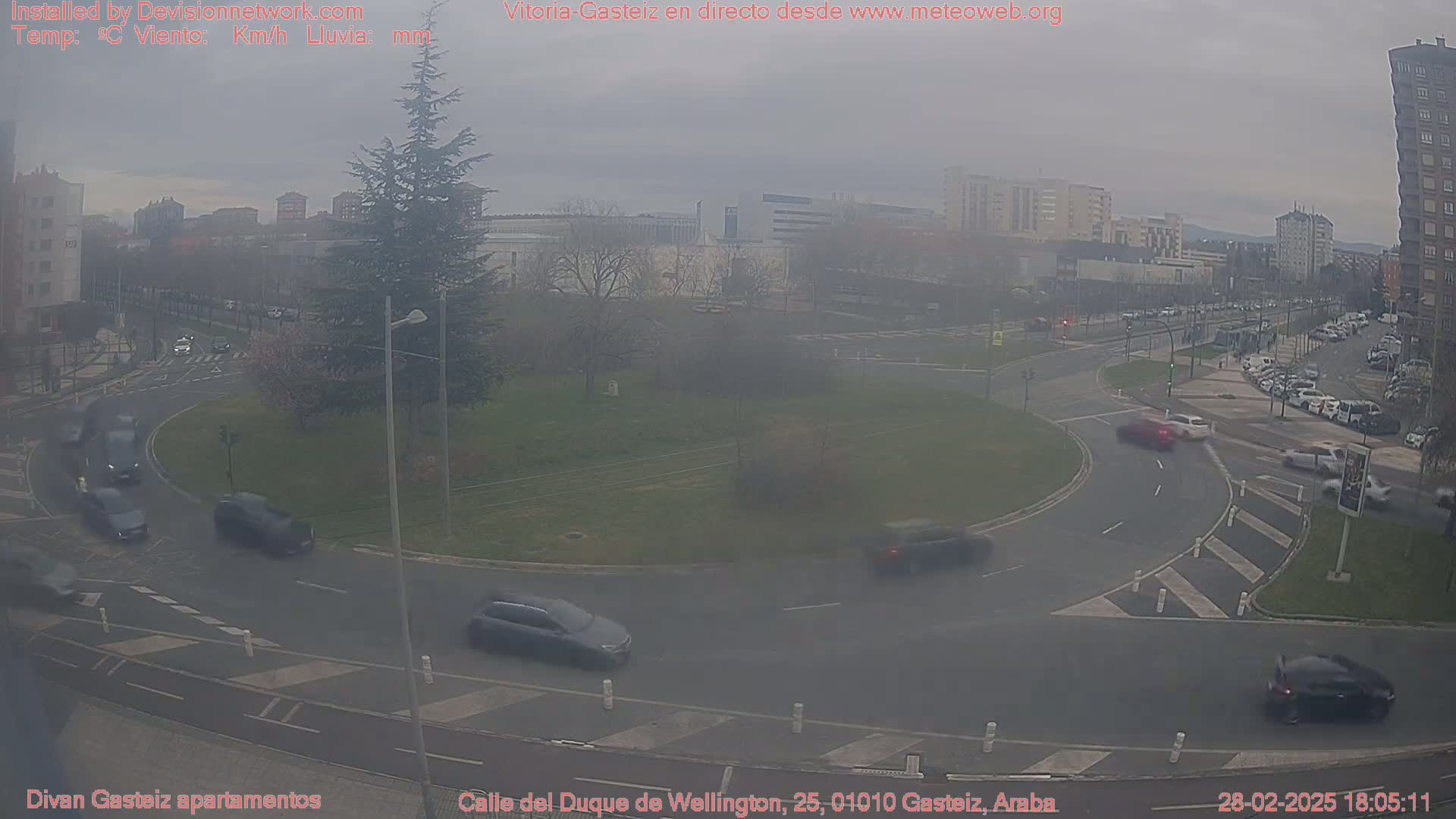 A hazy, overcast day reveals a roundabout with several cars navigating it, surrounded by buildings and trees.