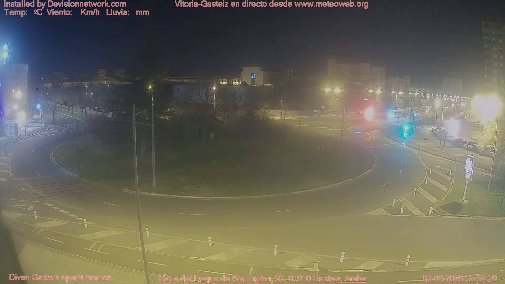 A nighttime view of a roundabout and surrounding streets with several vehicles present, under clear skies.