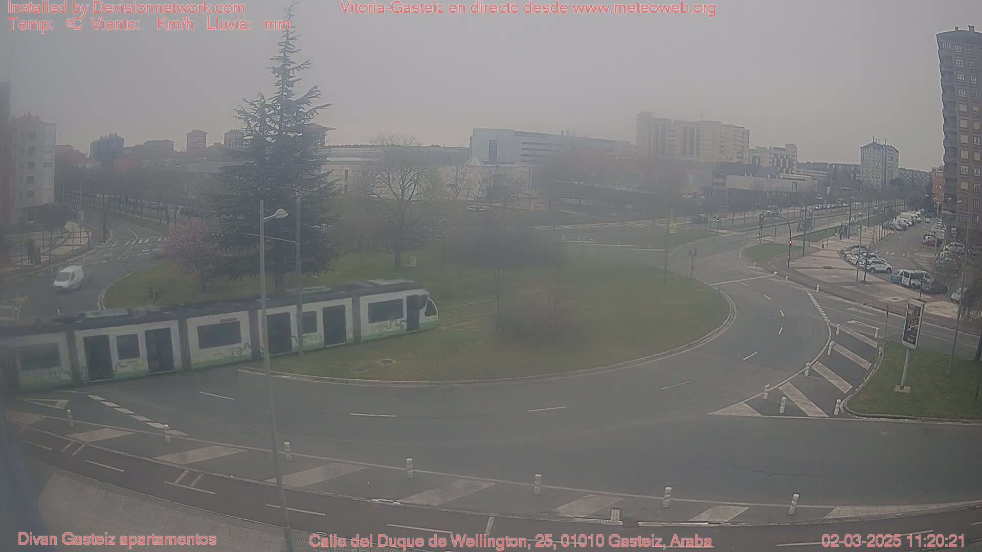 A hazy view of a city intersection with a tram stopped at a corner, several cars parked along the sides of the road, and a few cars driving.