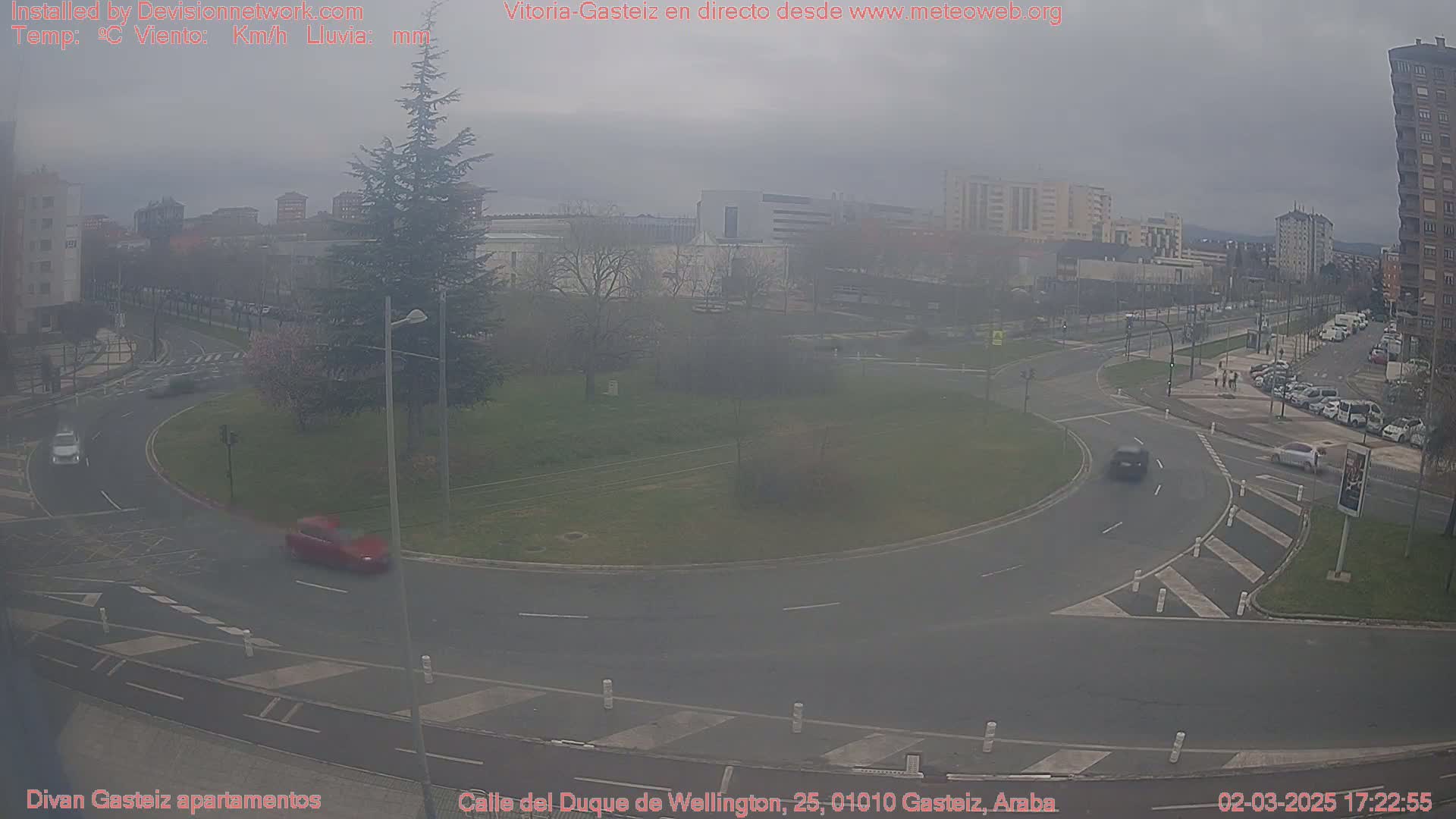 A hazy, overcast day reveals a roundabout in a city with cars driving around it and parked cars along a nearby street.